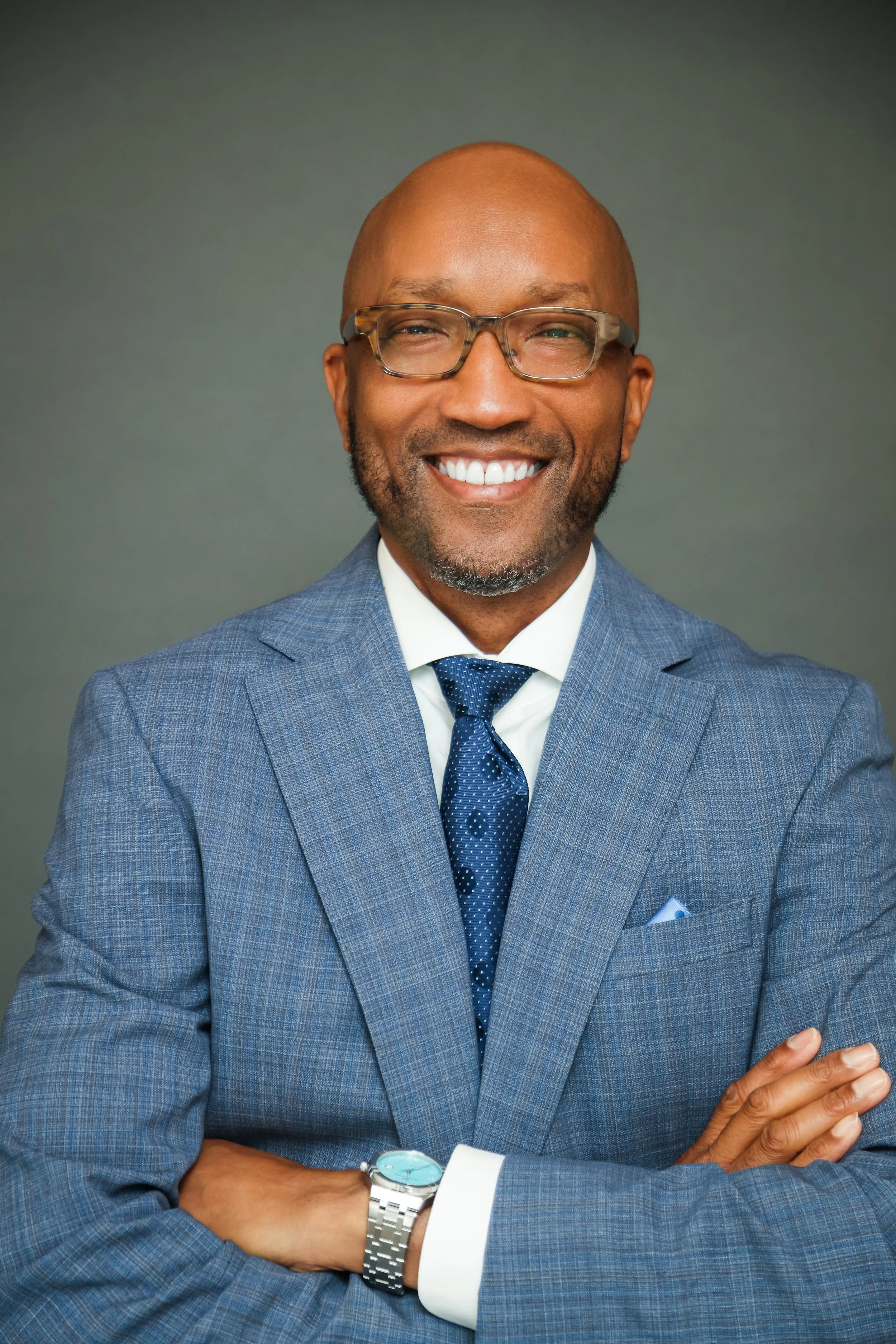 Close-up portrait of a smiling African American man wearing glasses, a blue suit, white shirt, and dark blue tie, with a watch on his left wrist, against a gray background.