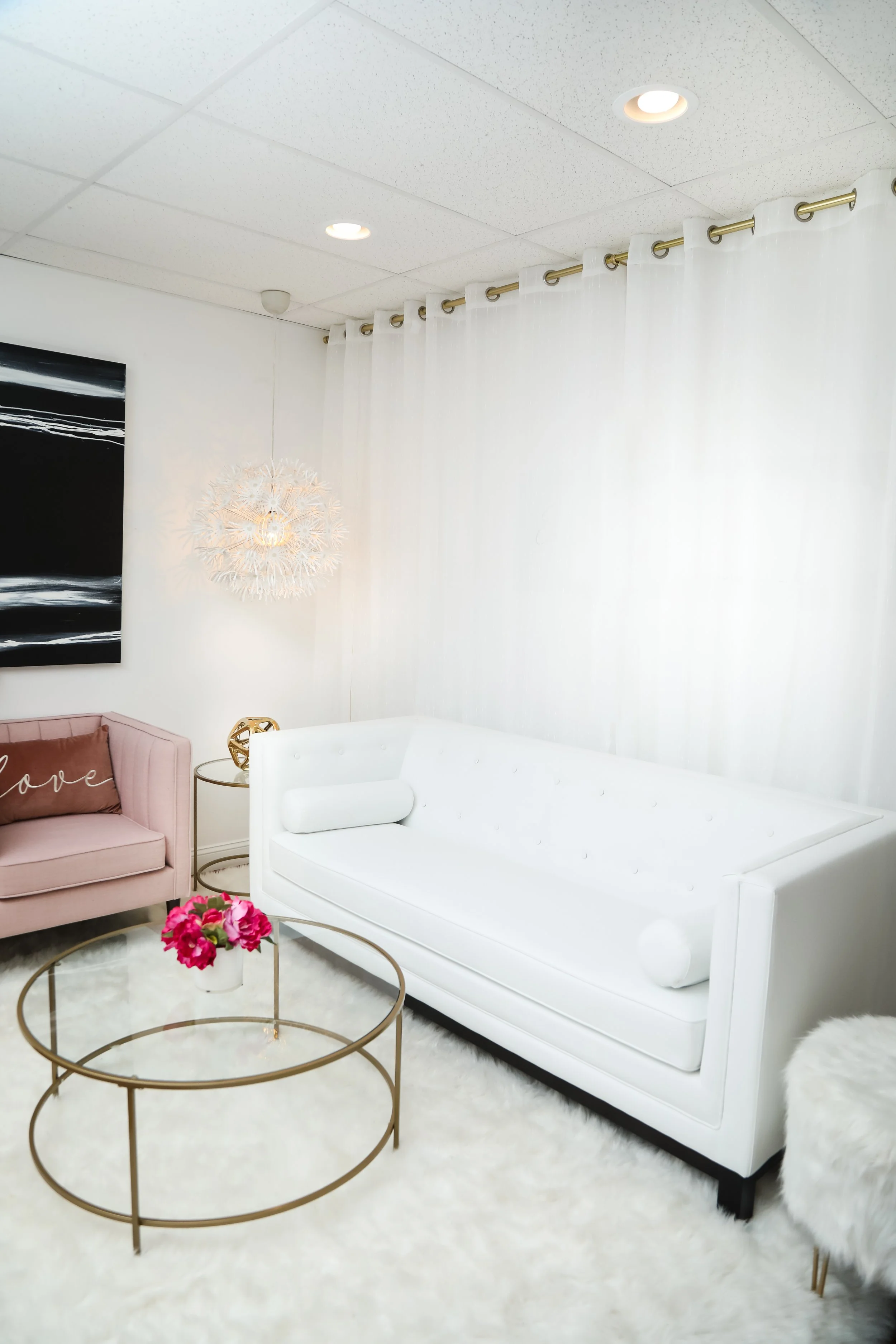 A modern, bright living room with white walls, white ceiling, and white furniture, including a sofa and a small white bed, accentuated with a pink pillow and pink flowers on a glass-topped coffee table, gold accents, black-and-white artwork, and a white shag rug.