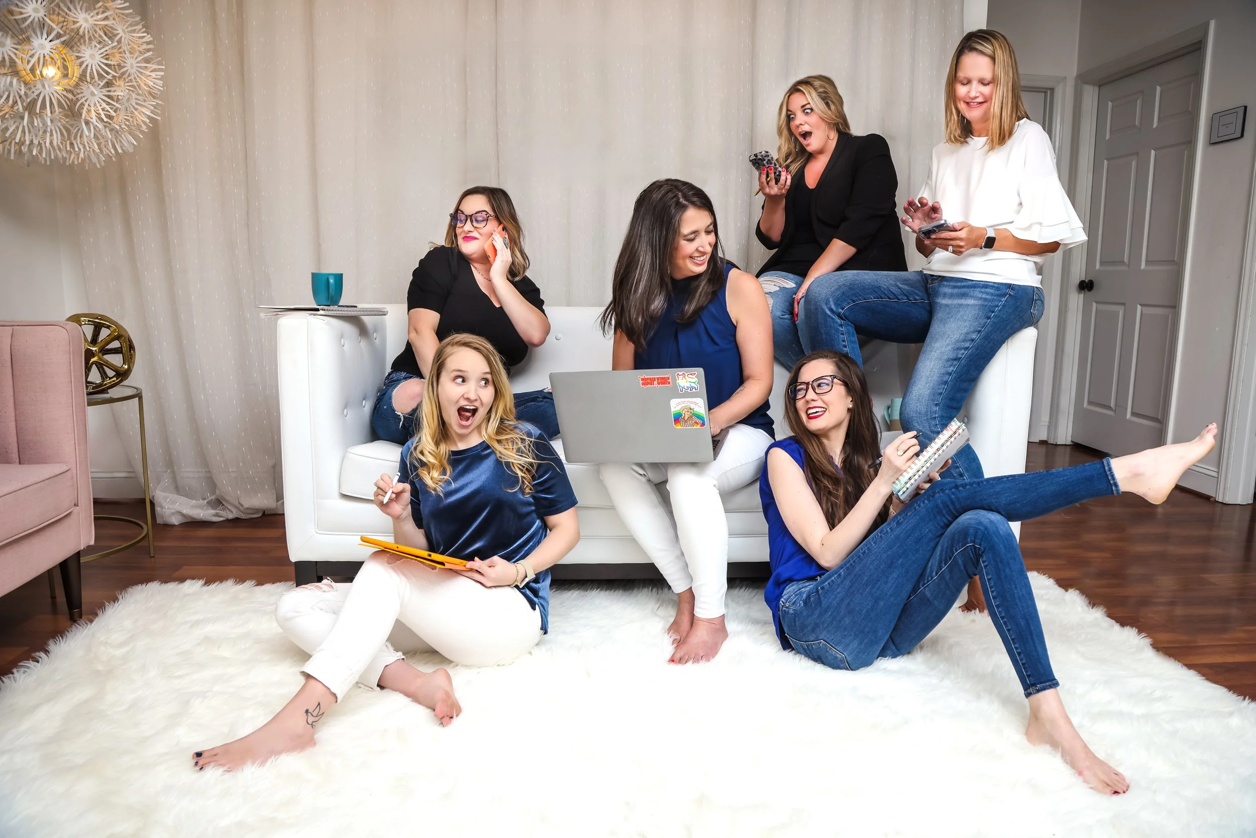 A group of six women relaxing and having fun together in a living room, sitting on and around a white sofa, with a wooden floor and a plush white rug; some are smiling, talking, and using their phones or a laptop.