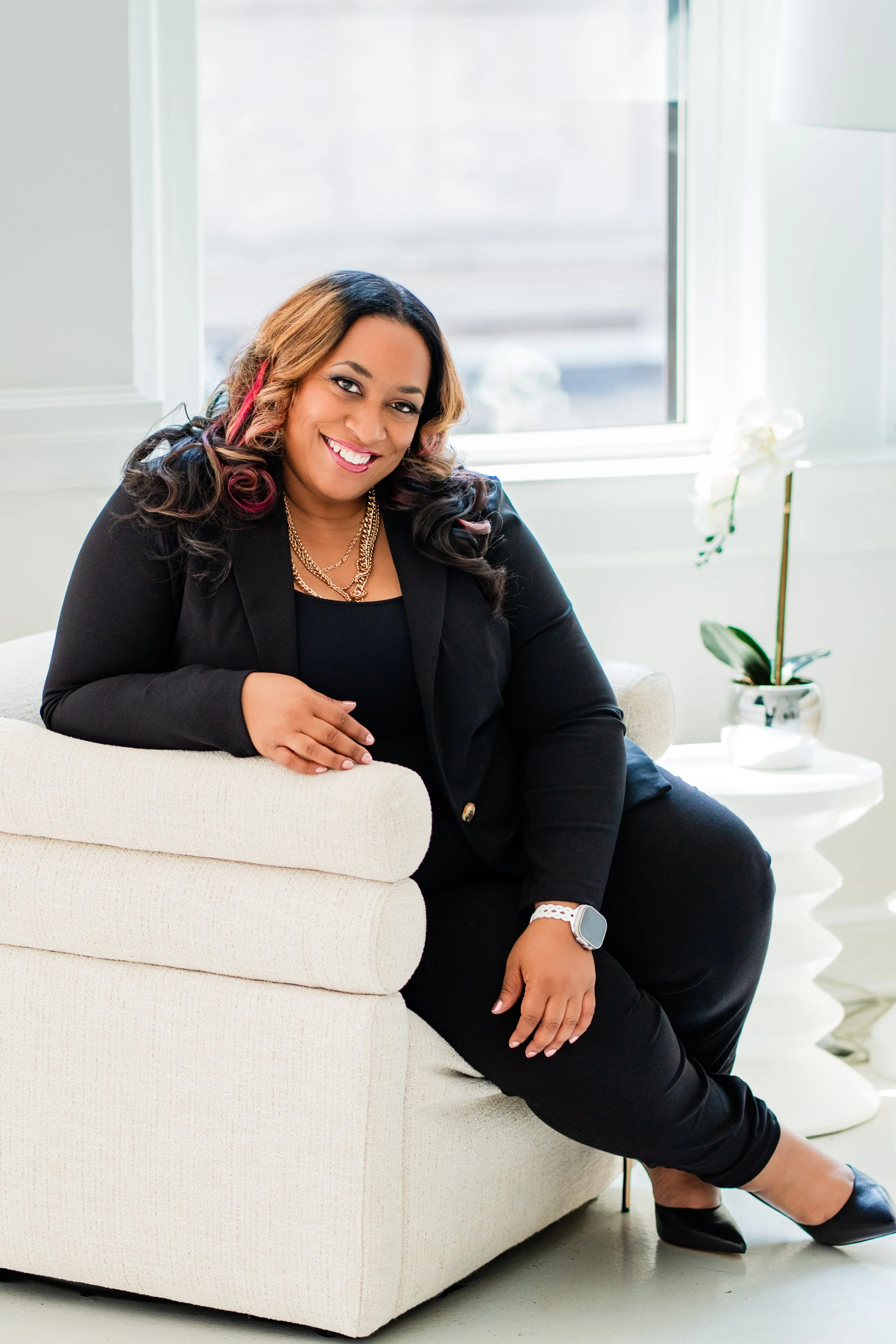 A woman with long wavy hair, wearing a black blazer and black pants, smiling and sitting on a beige sofa in a bright room with a window and a white orchid plant.