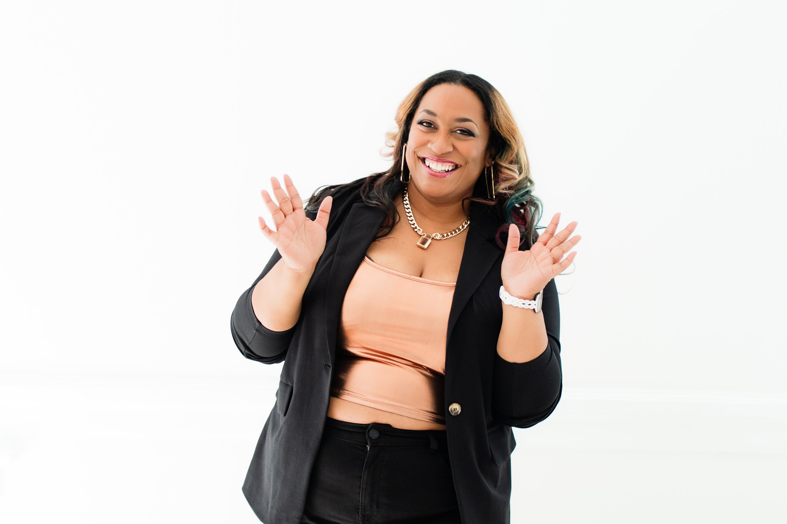 Smiling woman with curly hair, wearing a black blazer, light pink top, and gold jewelry, waving and looking at the camera against a white background.