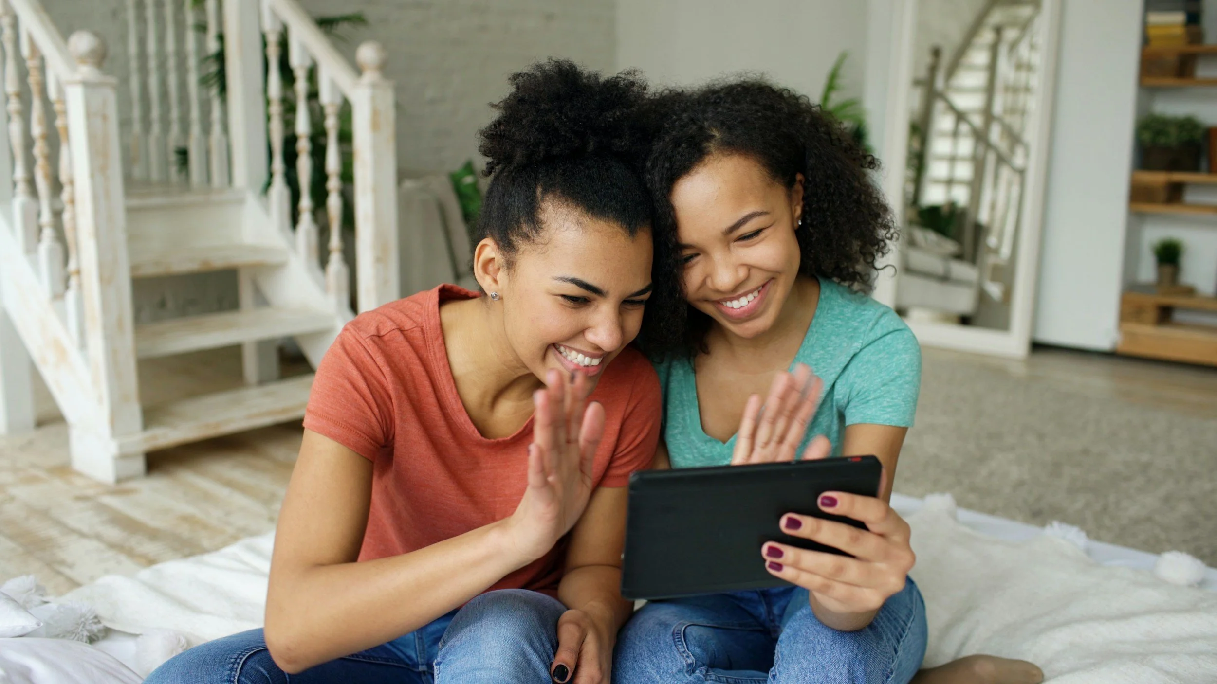 Two young women sitting on a bed, smiling and looking at a tablet together in a cozy, well-lit room.