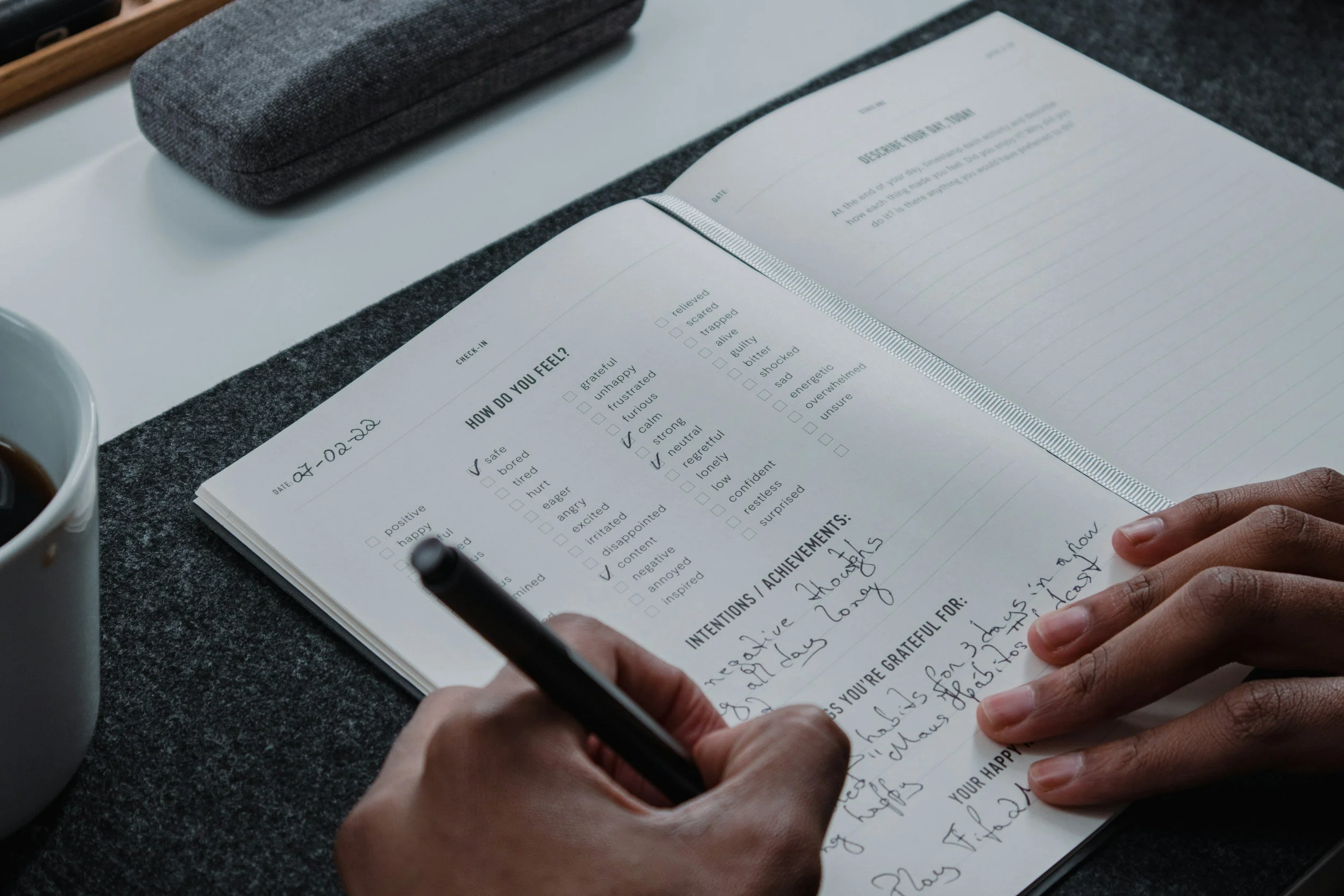 A person writes in a notebook with a black pen, with a gray case and a white mug nearby. The notebook is open to a page with checklist options and handwritten notes.
