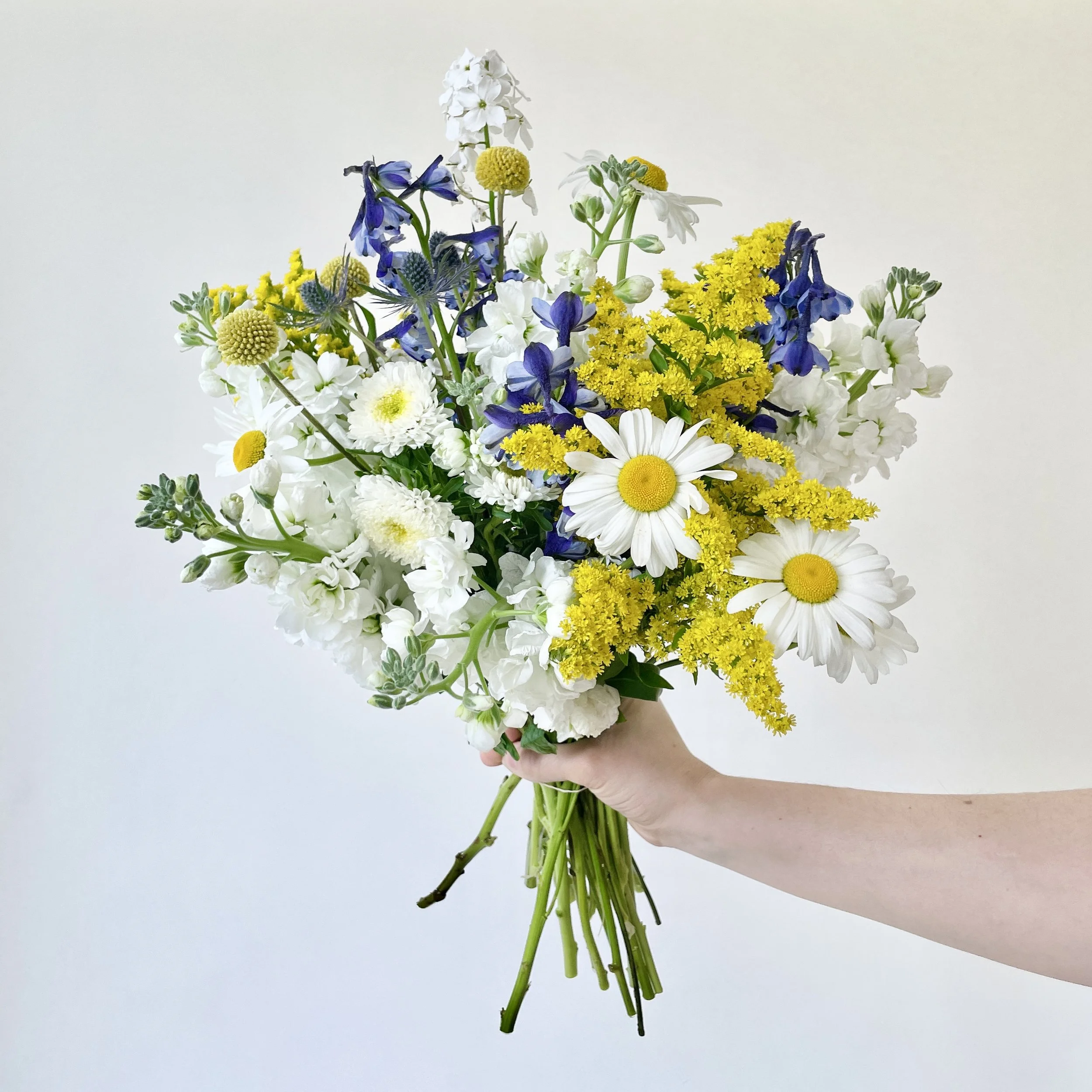 A bouquet of mixed white, yellow, purple, and green flowers held in a hand against a plain white background.