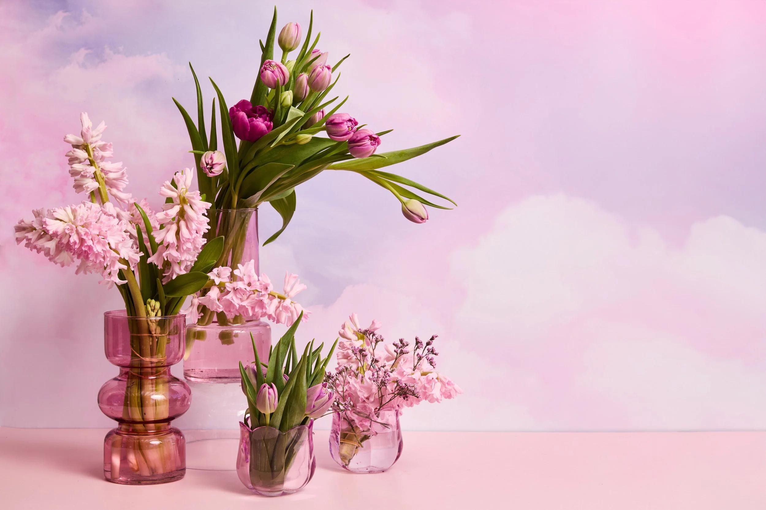 Several pink glass vases holding pink and purple tulips and hyacinths, arranged on a pink surface against a pink and purple sky background.