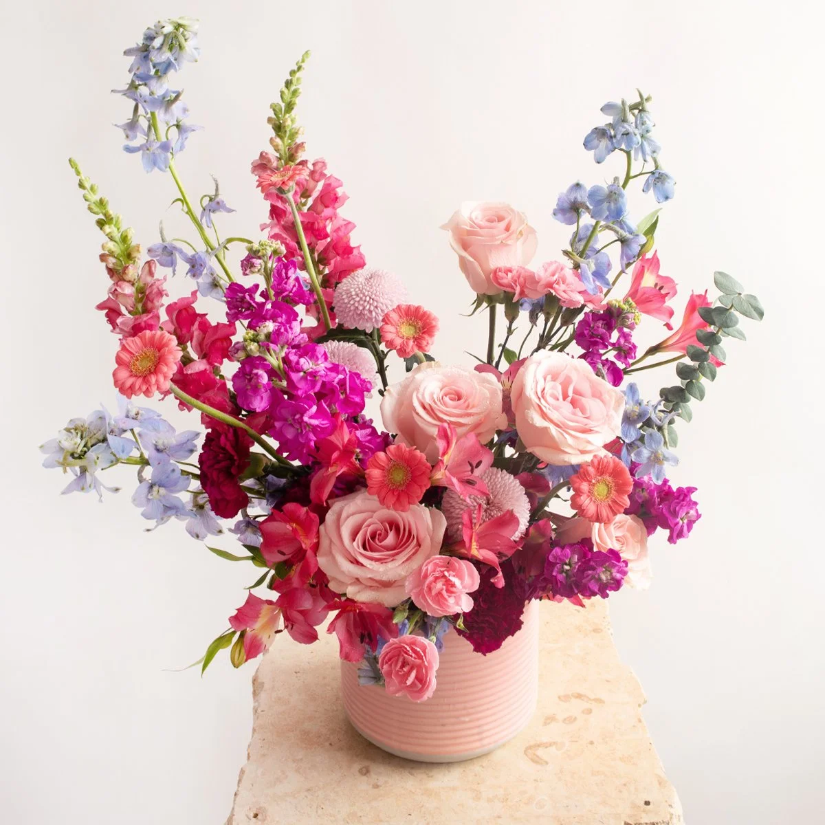 A pastel pink vase filled with a colorful bouquet of pink, purple, and blue flowers, including roses and other mixed blooms, placed on a beige surface against a plain white background.