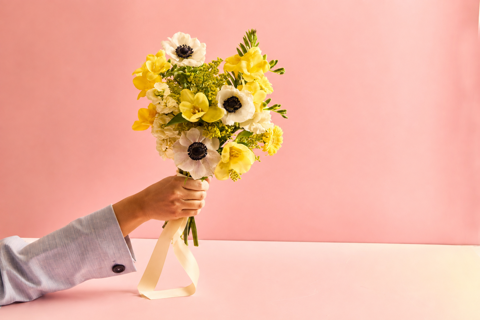 Hand holding a bouquet of yellow and white flowers against a pink background.