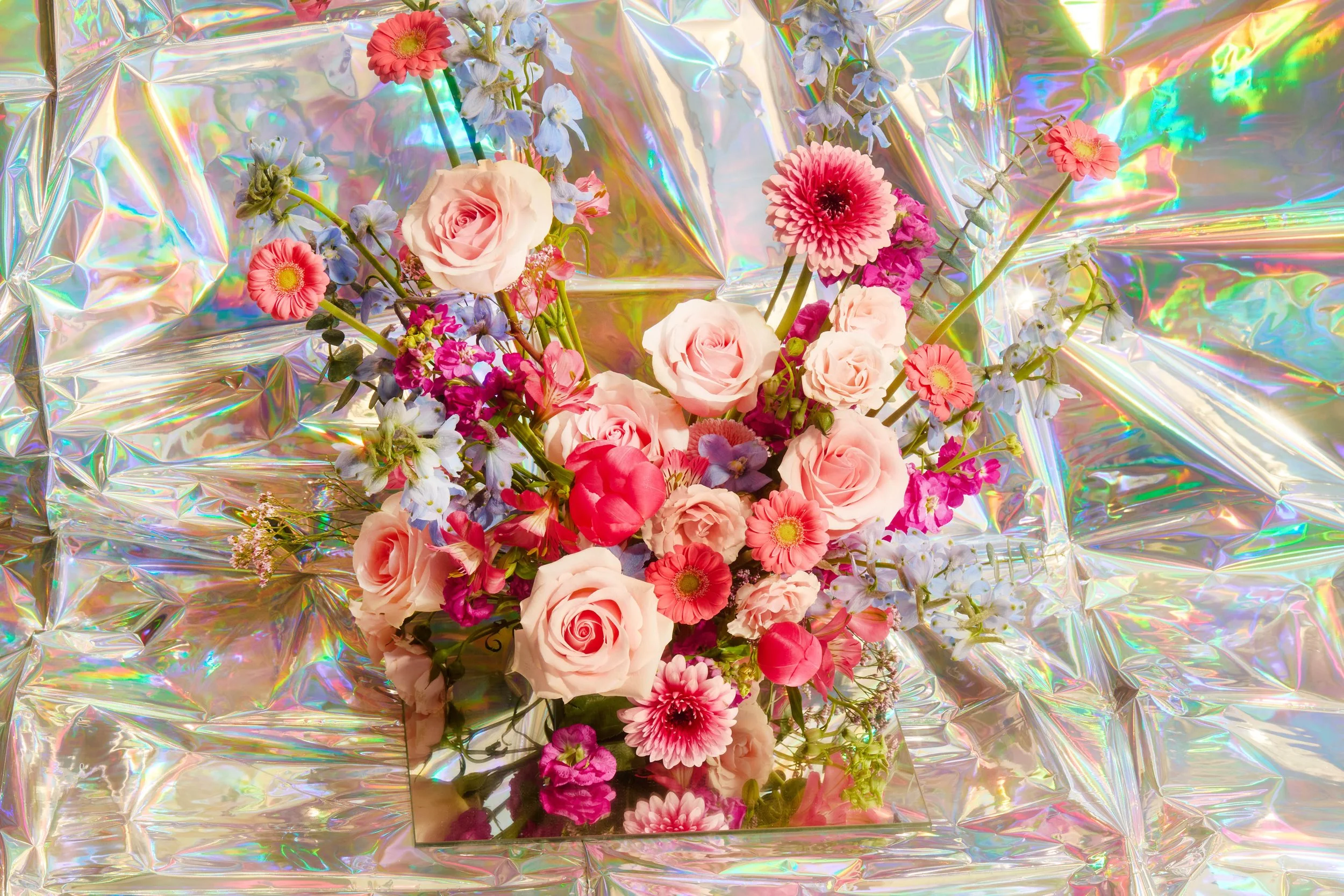 A colorful floral arrangement with pink roses, pink carnations, white and blue flowers, and greenery, placed on iridescent, holographic wrapping paper.