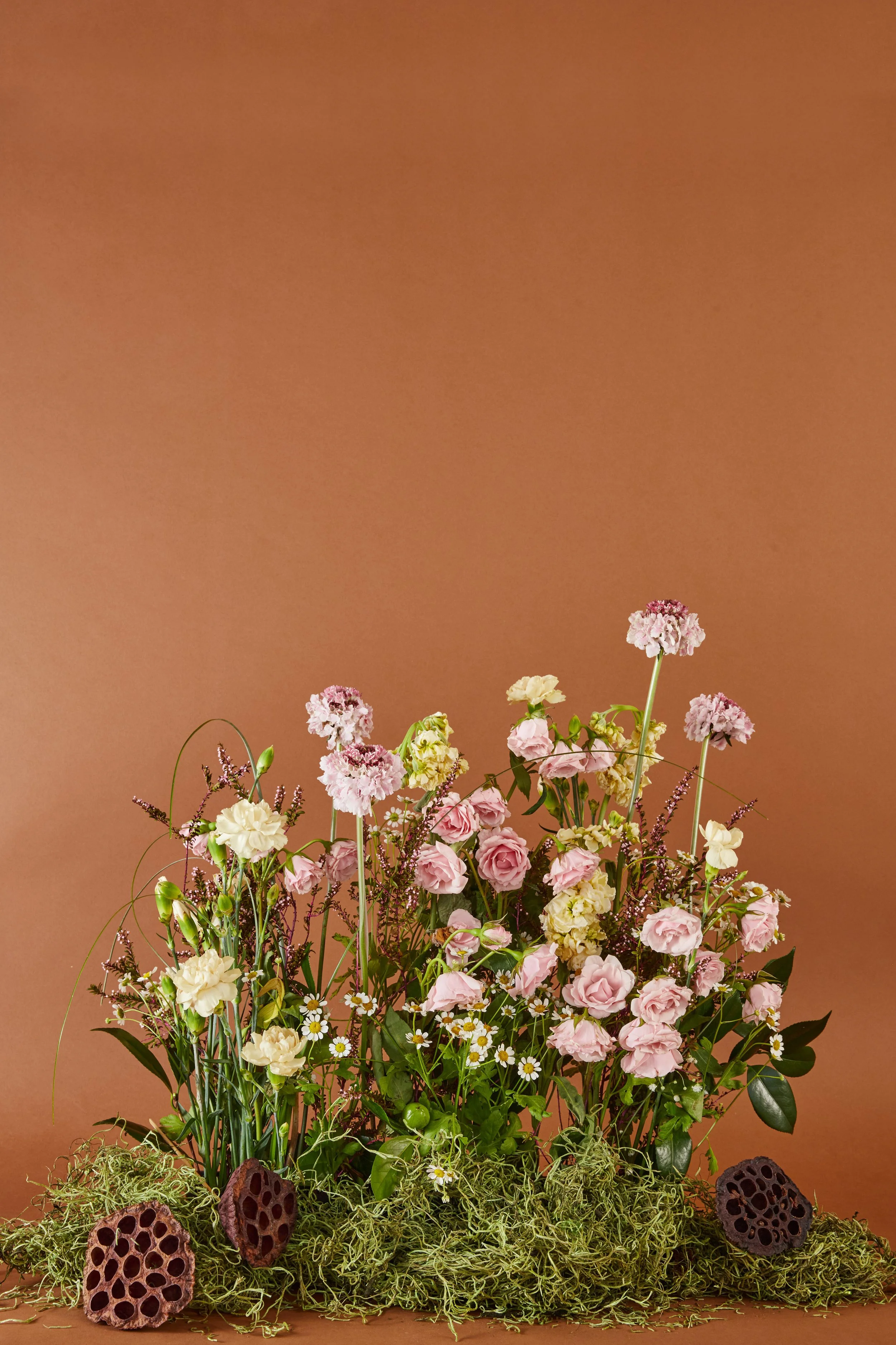 A floral arrangement with pink, white, and purple flowers, including roses and daisies, mixed with green foliage and decorated with dried lotus seed pods, set against a warm brown background.
