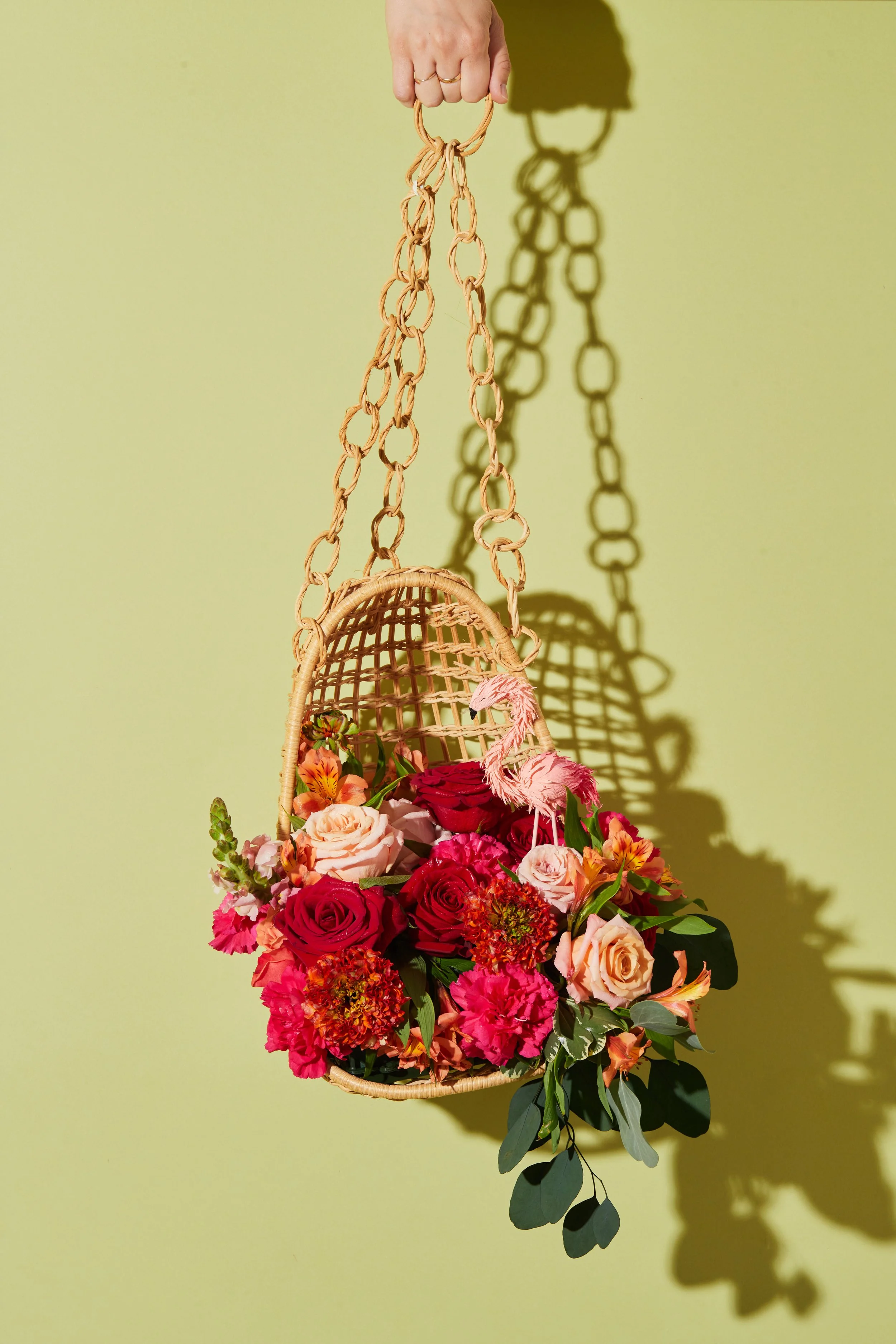 Hanging floral arrangement in a wicker basket with pink, red, and peach flowers, against a pale yellow wall.