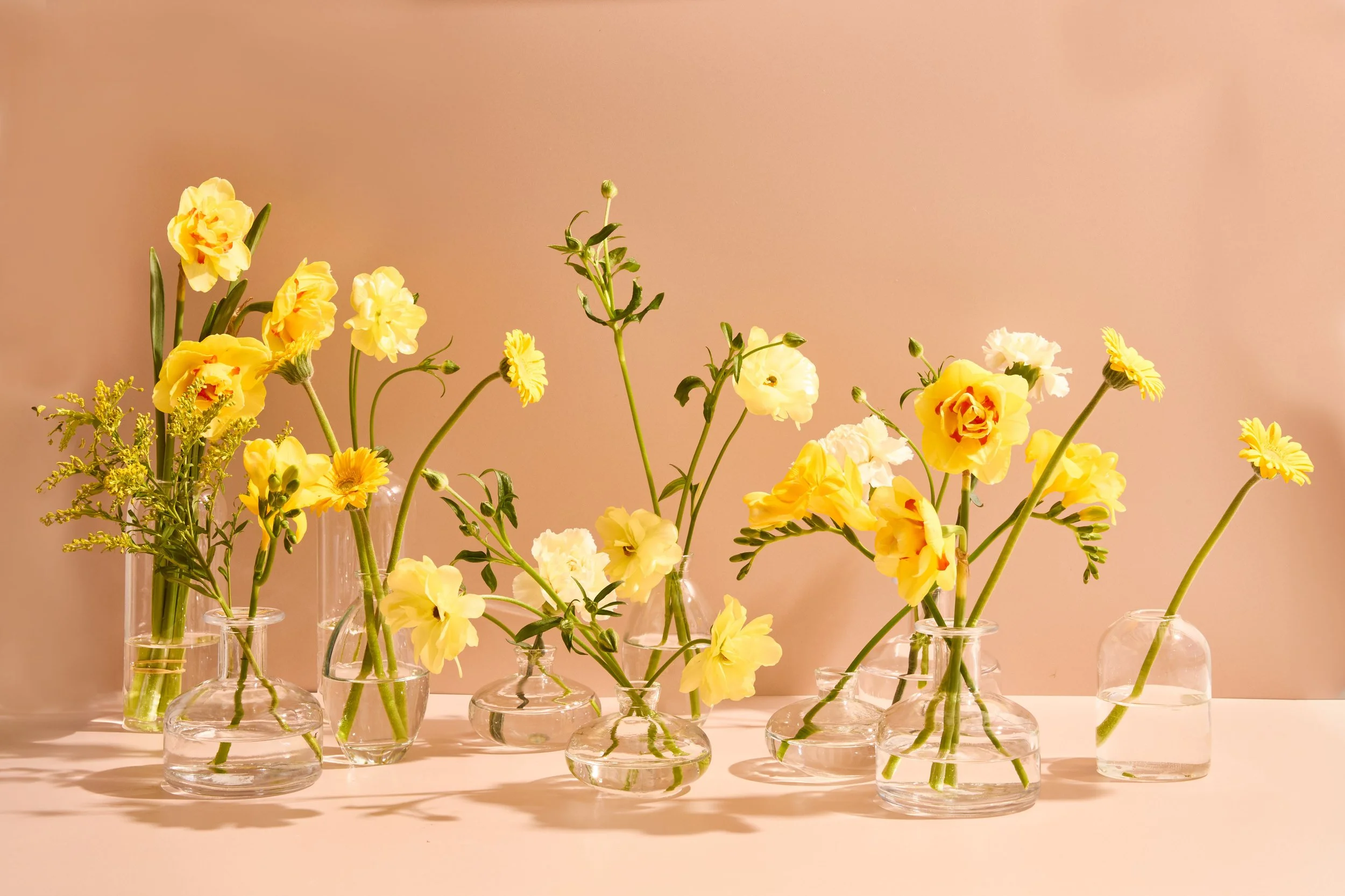 Various yellow and white flowers arranged in small glass vases on a light-colored surface with a plain beige background.