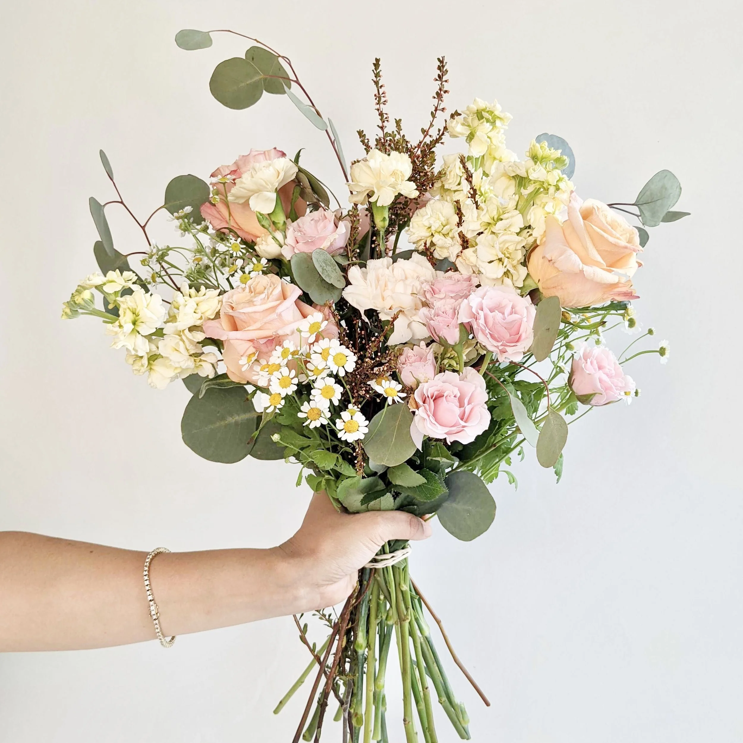 Person holding a bouquet of mixed flowers including pink roses, cream hydrangeas, small white daisies, and greenery against a plain light background.