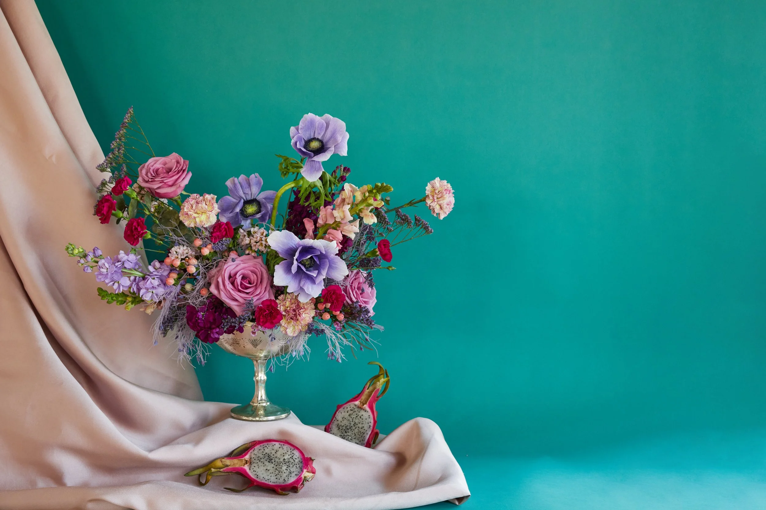 A bouquet of mixed flowers in a glass vase, placed on a satin cloth, with dragon fruit slices on the cloth and a teal background.