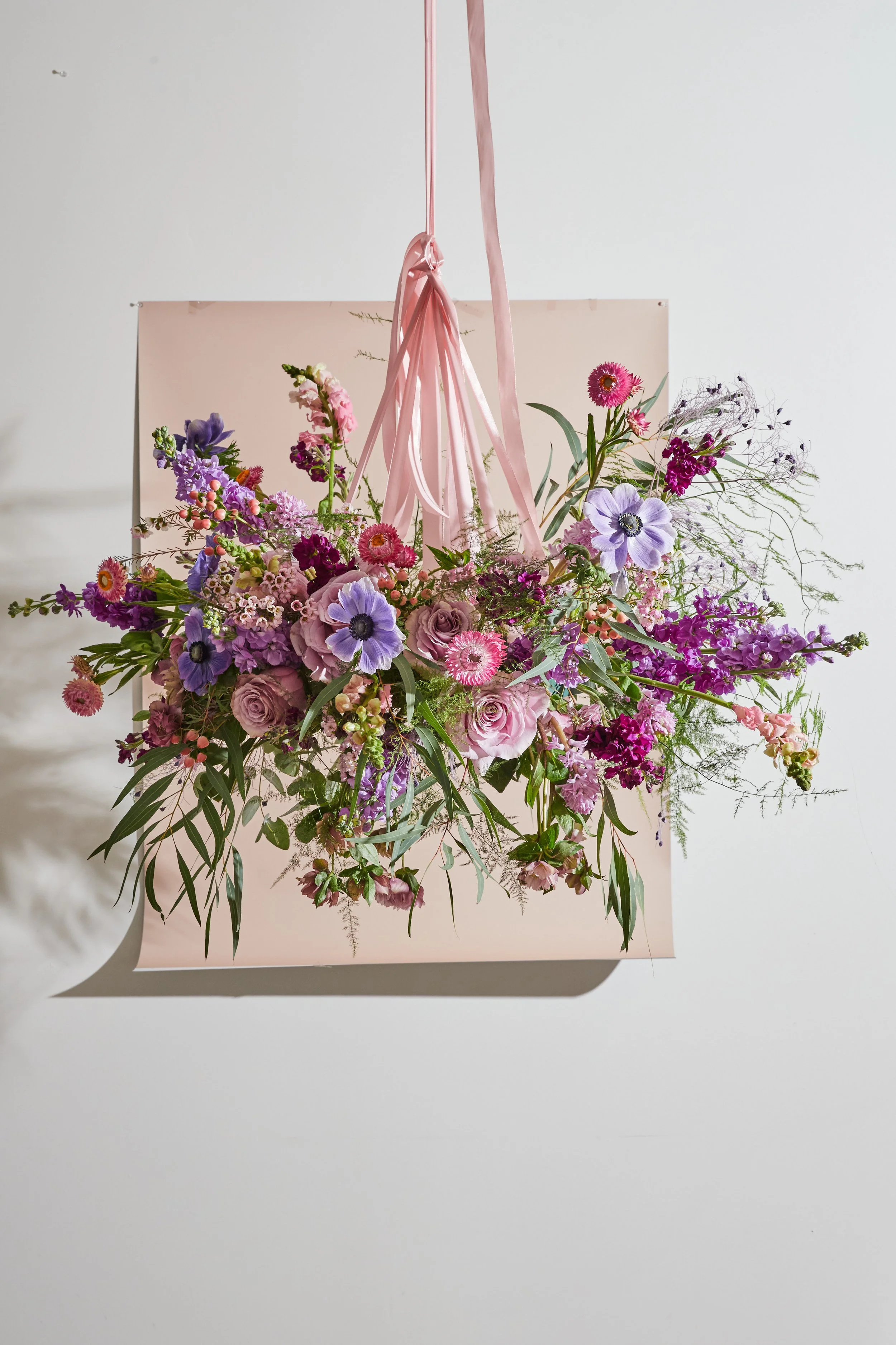 A large bouquet of pink, purple, and lavender flowers, arranged with greenery, hangs from a pink ribbon, with a light pink background and a white wall.