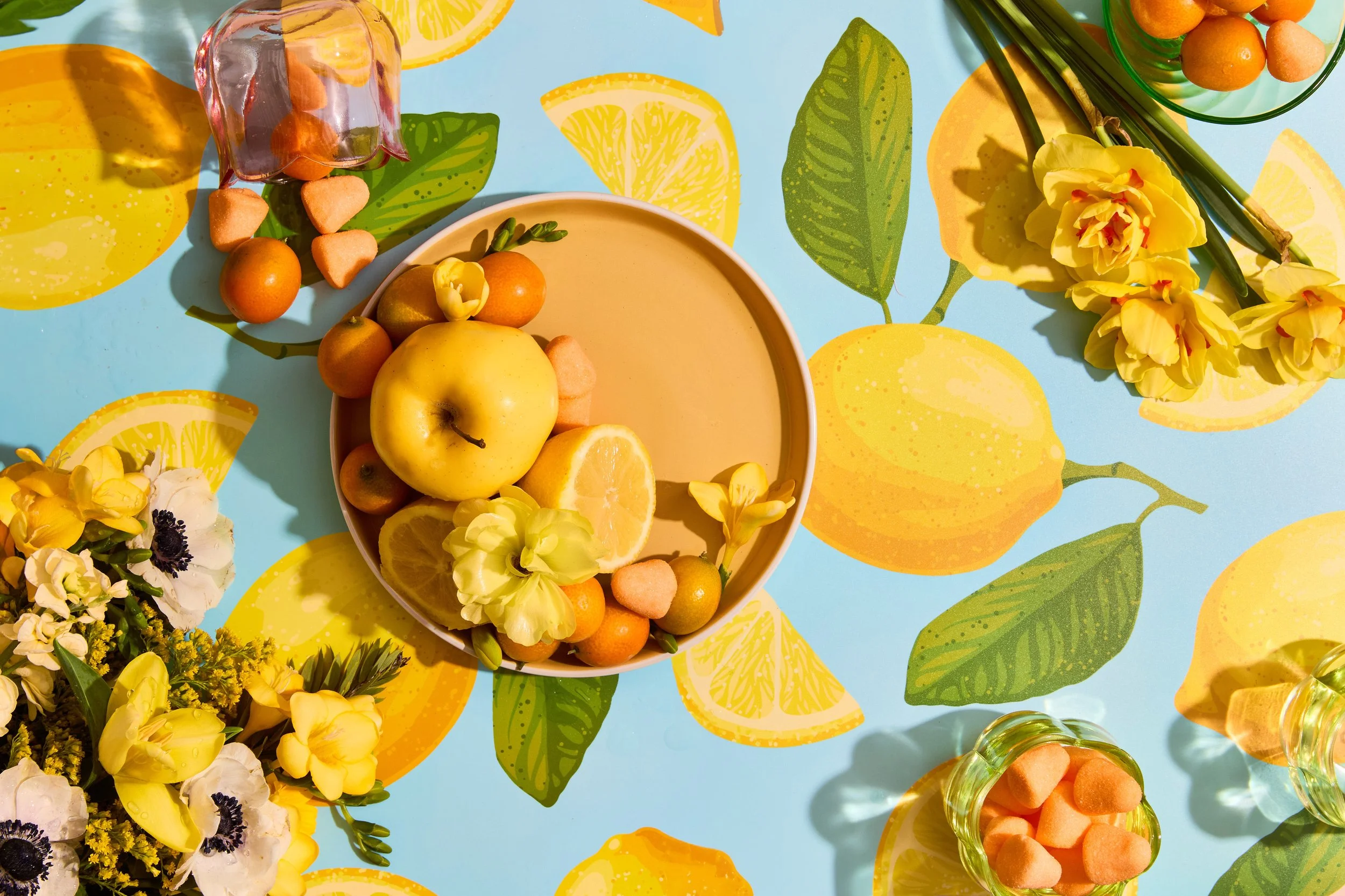 Table with yellow and citrus-themed decorations, featuring a bowl of lemons, oranges, and flowers, along with small orange heart-shaped candies and flower arrangements on a bright, lemon-patterned tablecloth.