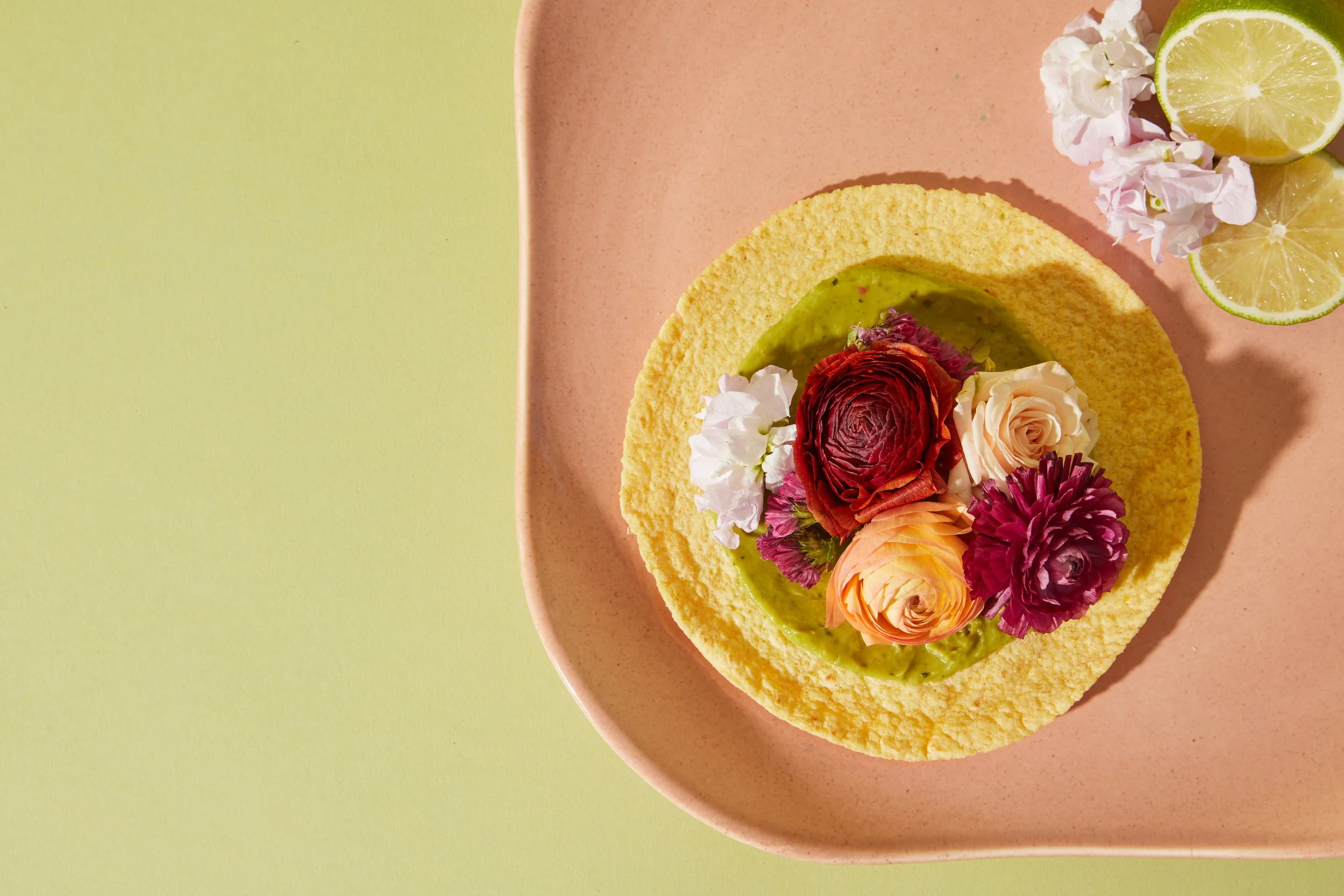 A plate with a taco shell topped with guacamole and colorful edible flowers, with lemon slices and white flowers on the side.