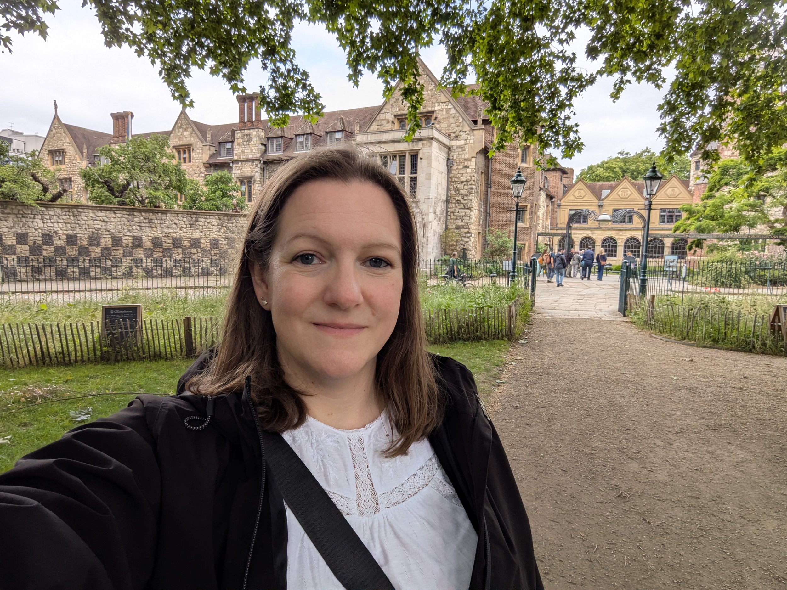 Elaine from Magpie Tours London , wearing a black jacket and white top, taking a selfie in front of historic stone and brick buildings with trees and a park in the background.