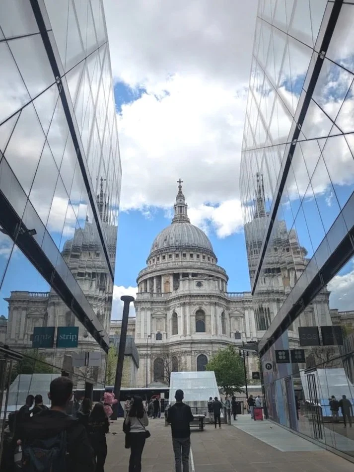 View of St. Paul's Cathedral in London, seen through modern glass buildings, with people walking in the foreground on a partly cloudy day.