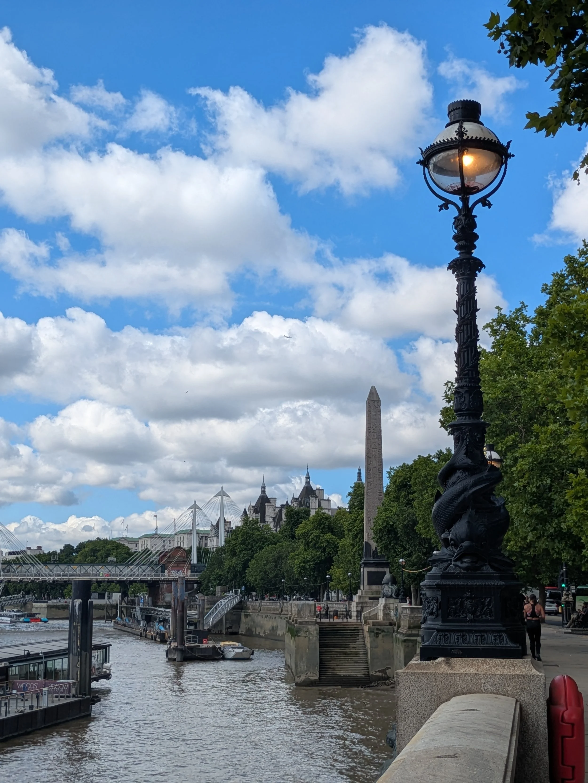 A scenic view of a river with a boat, a bridge, and historic buildings in the background under a partly cloudy sky. A black ornate street lamp is in the foreground, and there are trees and people walking along the riverbank.