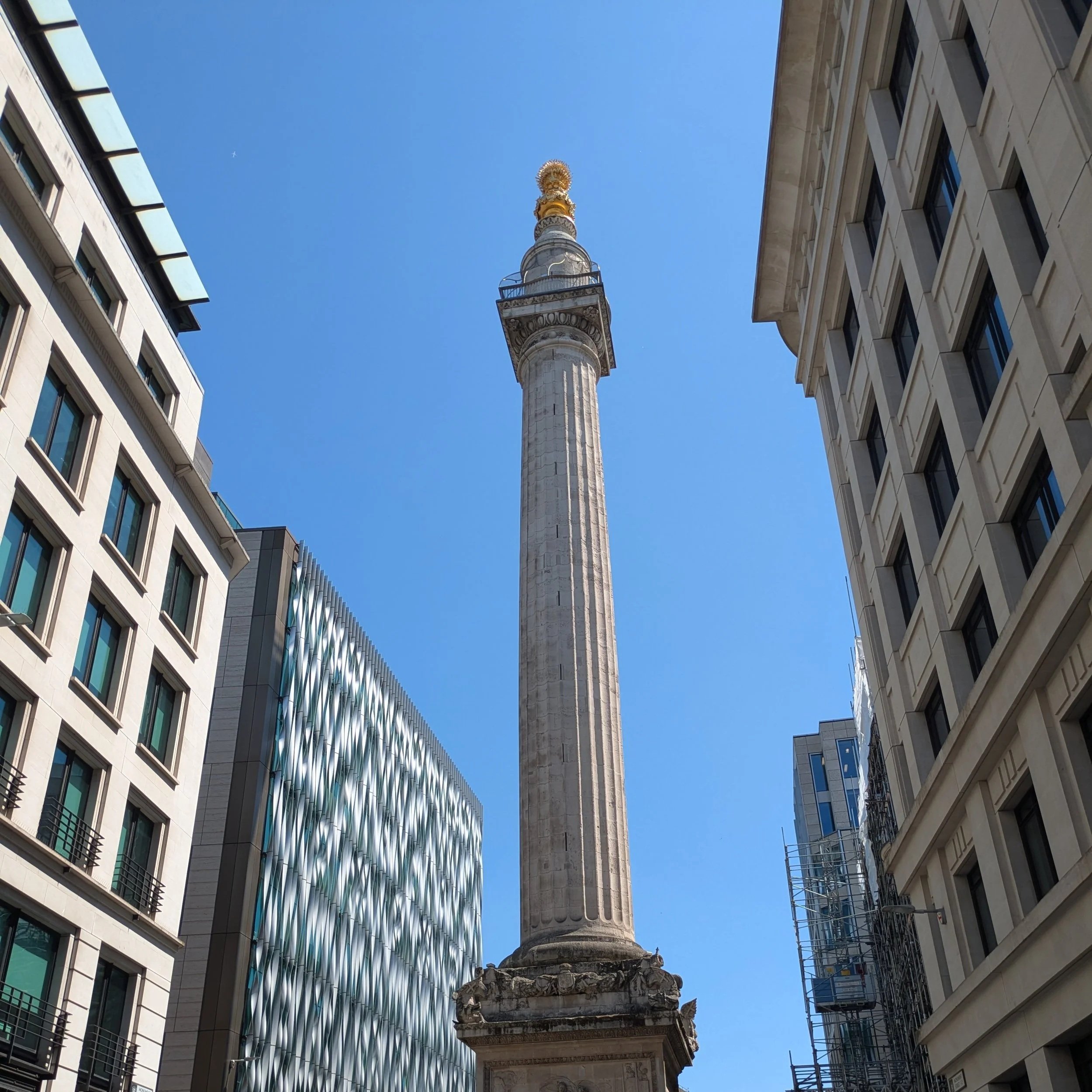 The Monument to the Great Fire of London. A tall decorative column in a city street, surrounded by modern office buildings with large windows, under a bright blue sky.