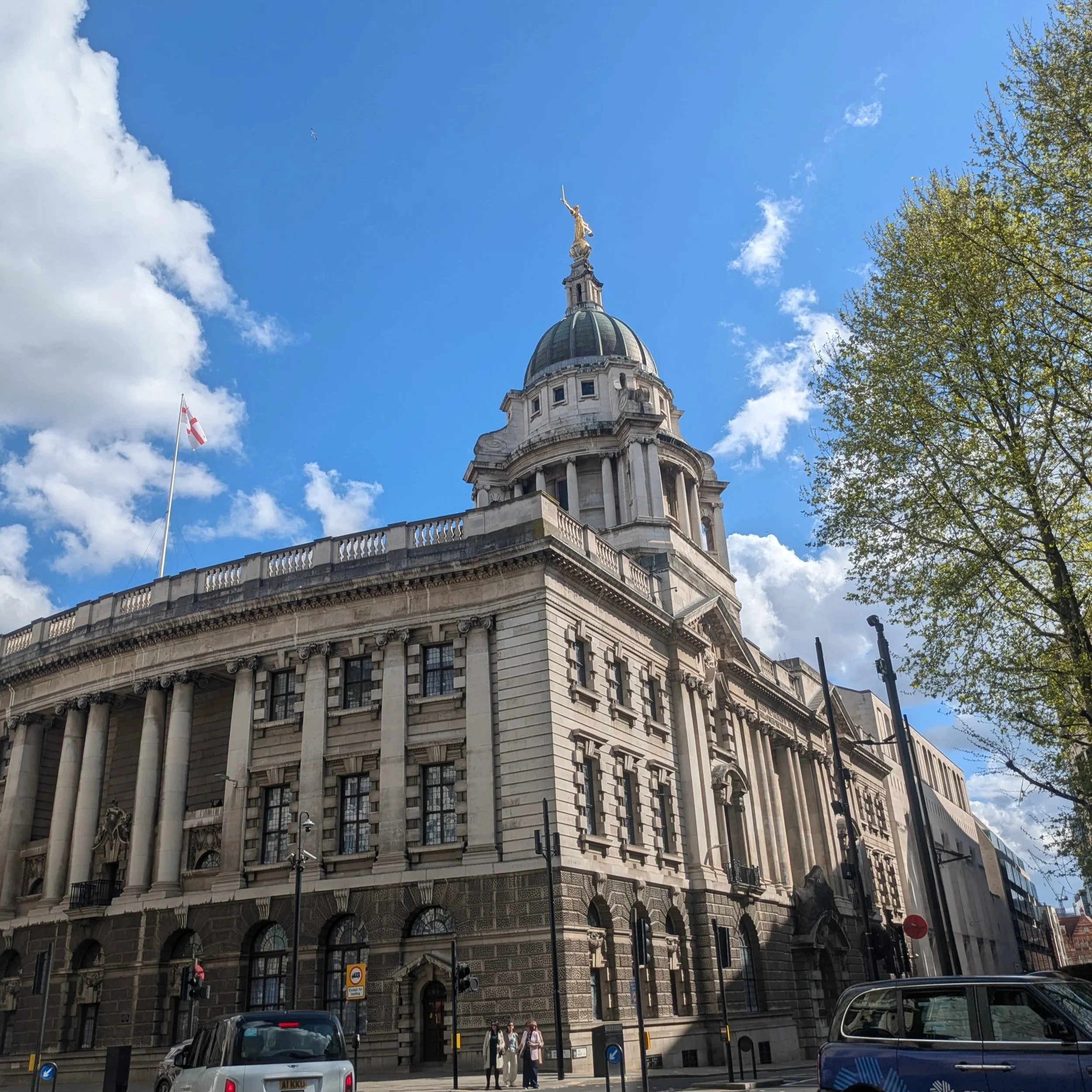 The Old Bailey, Central Criminal Court. A historic building with a rounded dome topped by a statue of a woman holding a sword, set against a blue sky with a few clouds. 