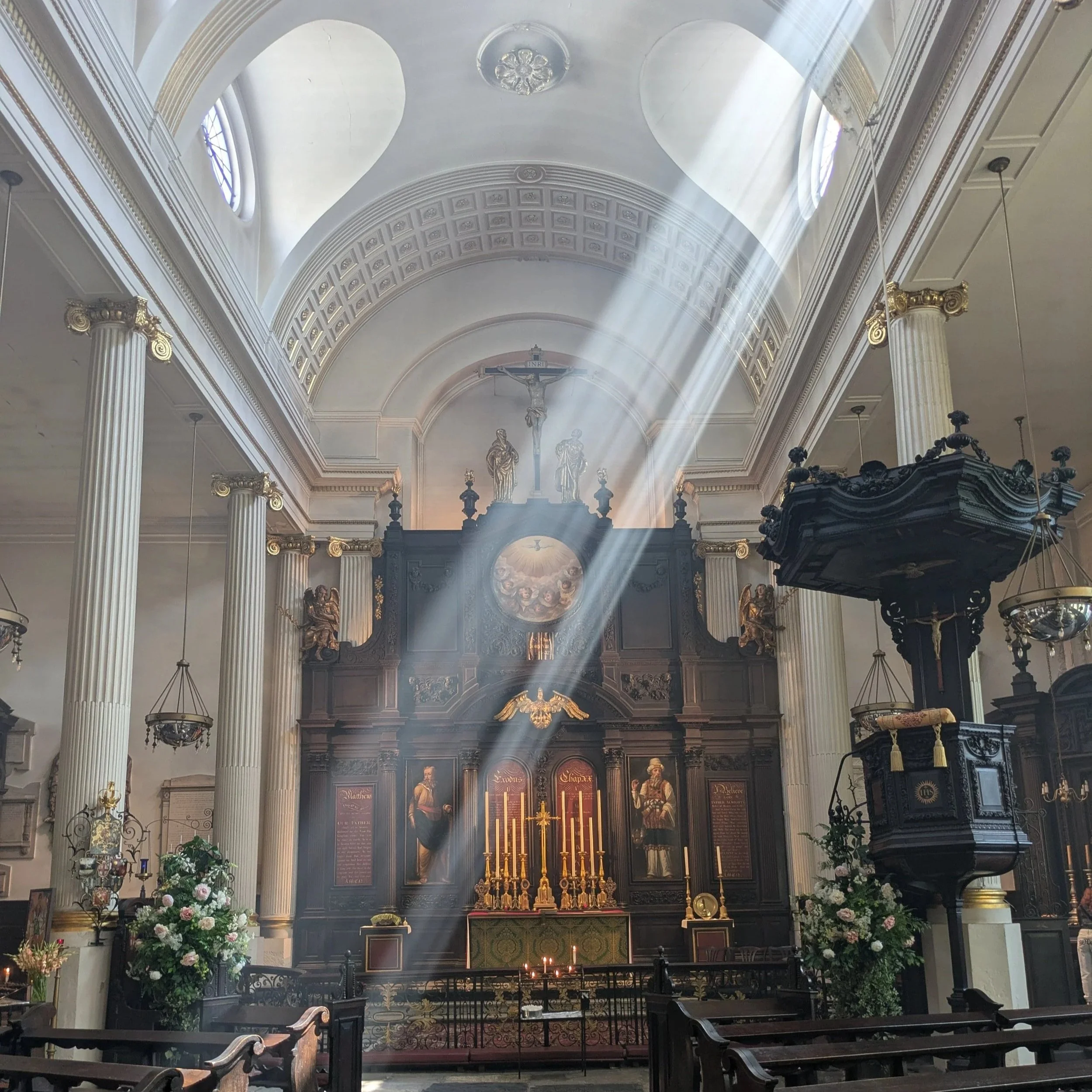Interior view of a church or cathedral with high arched ceilings, sunlight streaming through windows, columns with gold accents, religious statues, and an altar with candles and floral arrangements.