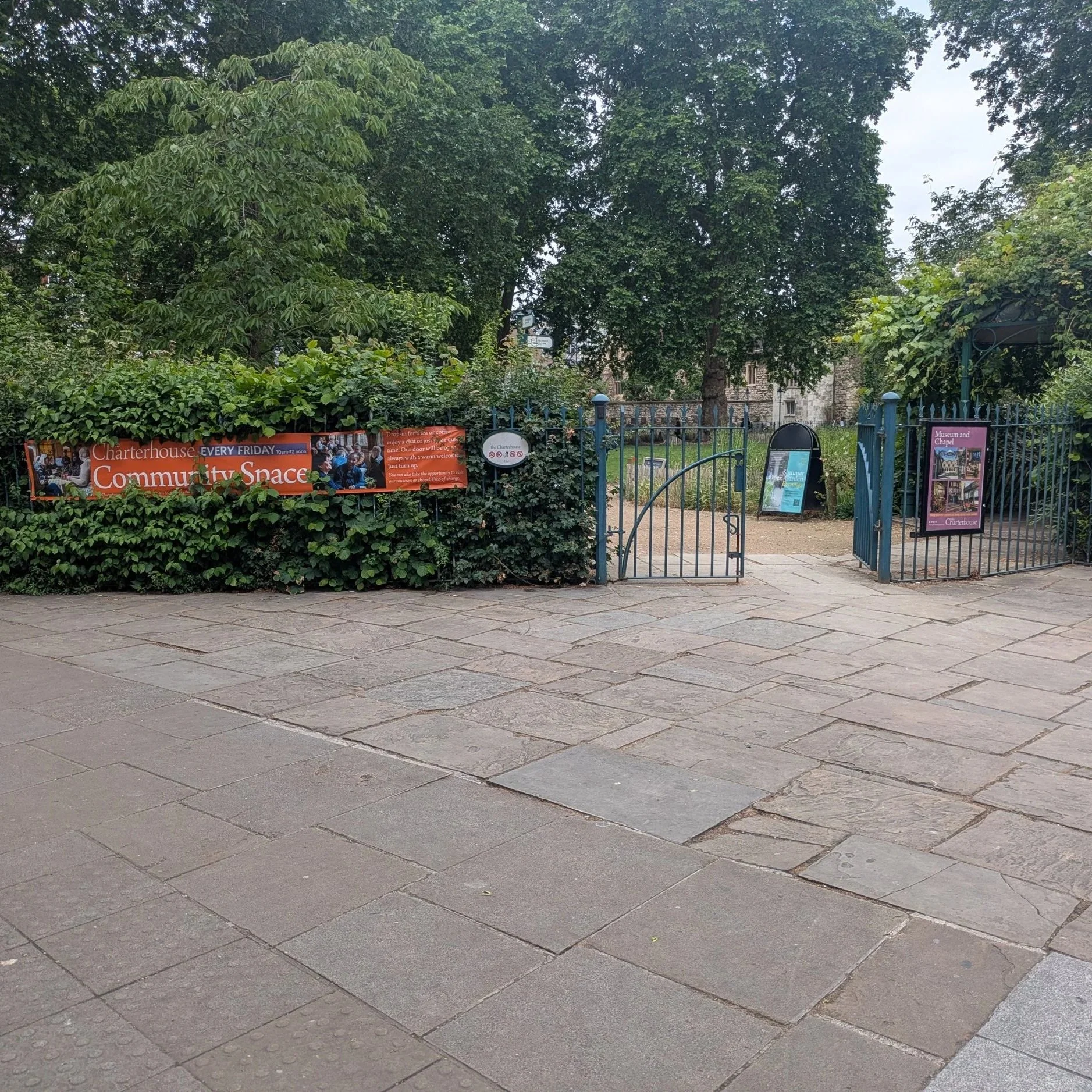 View of a park entrance with signs and informational posters, surrounded by green trees and foliage, a paved walkway in the foreground.