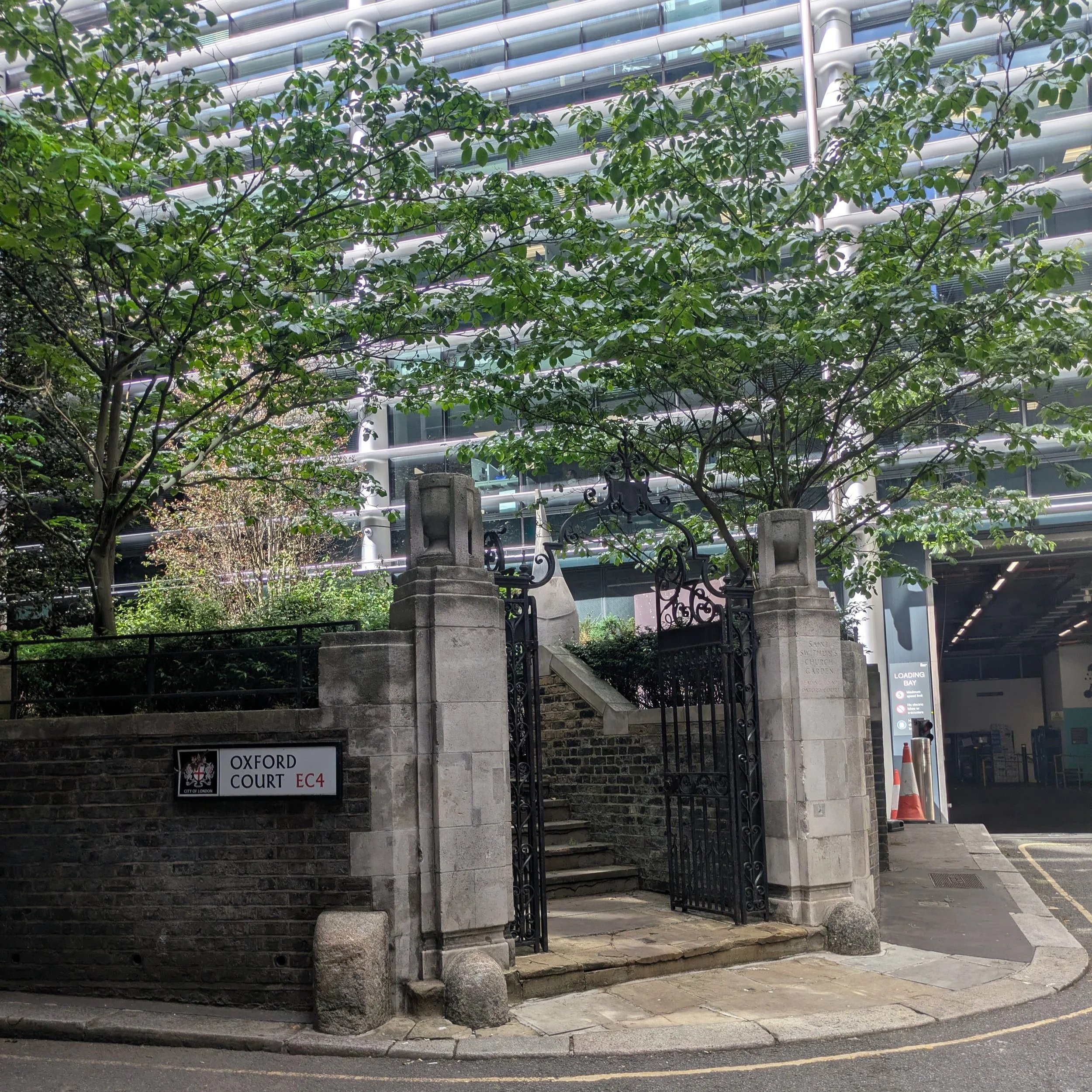 The image shows the entrance to Oxford Court, a building with a sign indicating its name. In front of the entrance, there are stone stairs, decorative metal gates, and stone pillars. Green trees partially obscure the view of the building, which has a