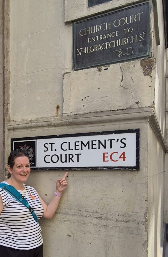 Elaine from Magpie Tours London pointing at a street sign that reads 'St. Clement's Court, EC4' on a historic building, with another sign above indicating the entrance to a church court on Gracechurch Street. Part of the Rhymes and Reasons tour.