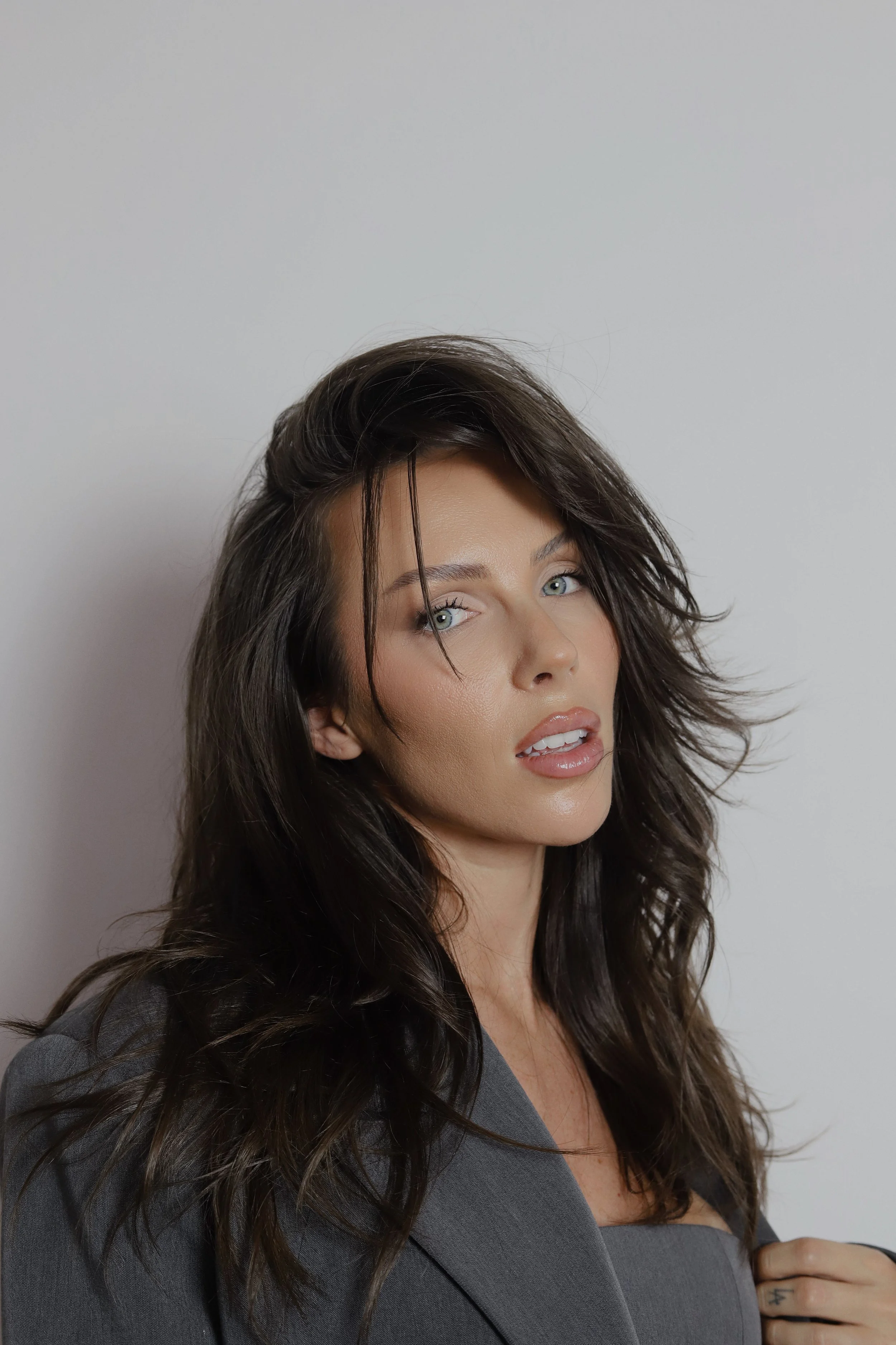 A woman with long, wavy dark hair, blue eyes, and light makeup wearing a gray blazer, posing against a plain white background.