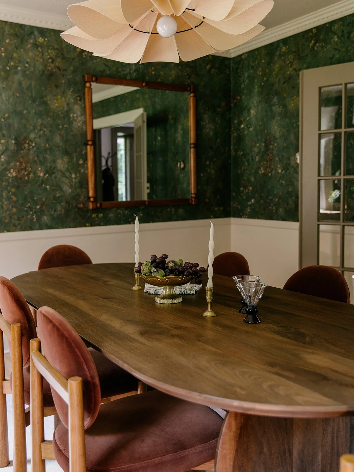 A dining room with a wooden oval table, pink velvet chairs, a bowl of grapes, two white twisted candles in gold holders, and a modern pendant light fixture. The wall is decorated with dark green textured wallpaper and a large mirror with a wooden frame.