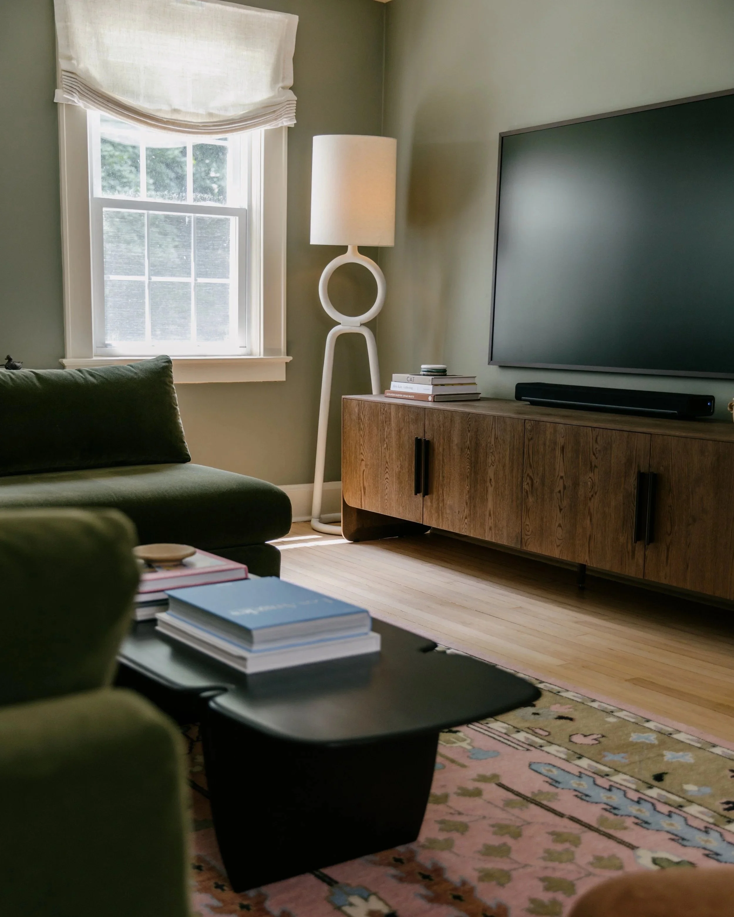 Living room with a window, white window frame, light-colored walls, a black and green chair, a wooden TV stand, a large flat-screen TV, a tall white floor lamp, a stack of books, a pink and blue patterned rug, and a patterned area rug.