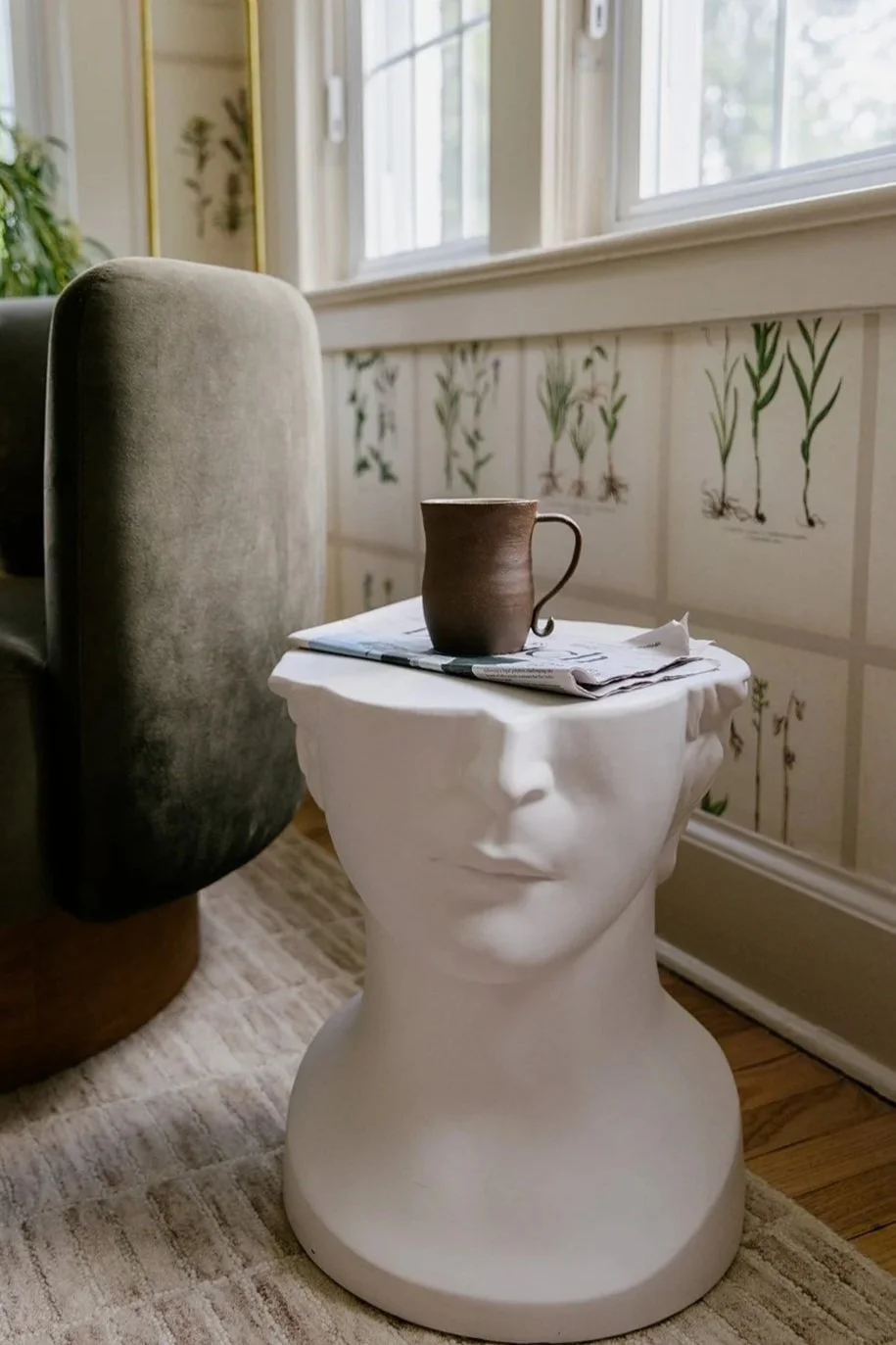 A white sculpture of a human head with a fabric-like top surface, used as a table, placed on a carpeted floor near a window. On top of the sculpture is a brown ceramic mug and a newspaper.