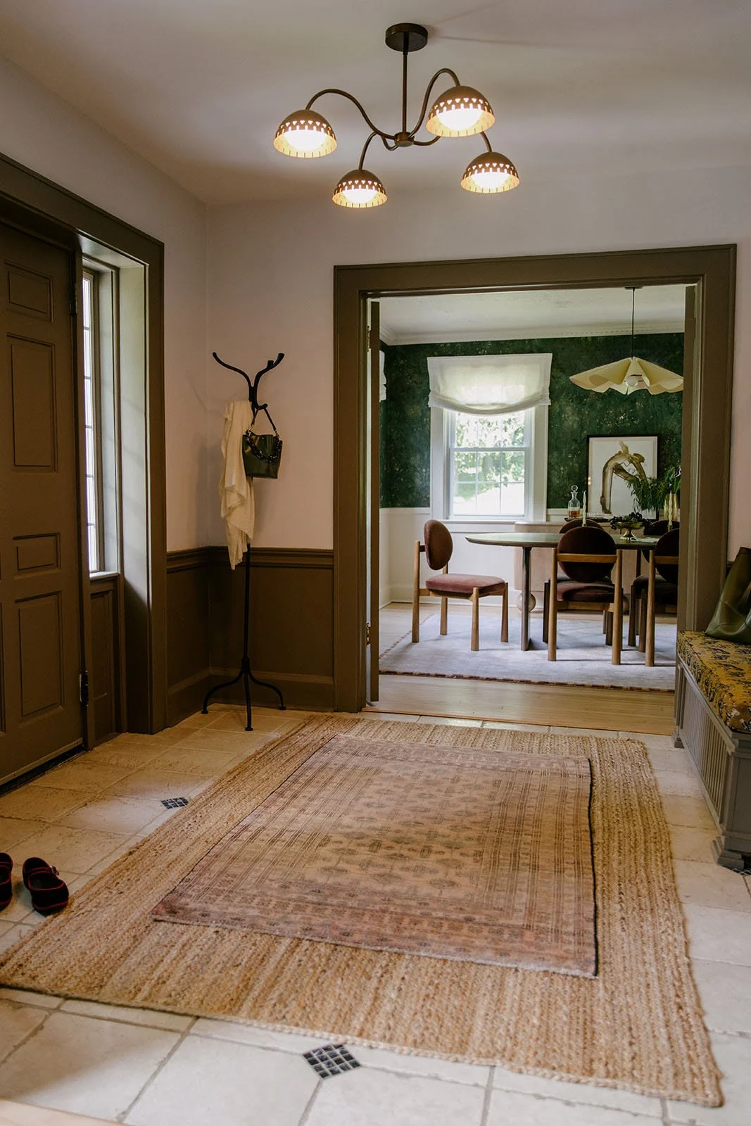 Interior view of an entryway with a rug on the tiled floor, a coat stand with a white towel and handbag, and a doorway leading to a dining room with a round table and four chairs. The dining room has a window with a white shade and a green patterned wall, and a chandelier hangs from the ceiling.