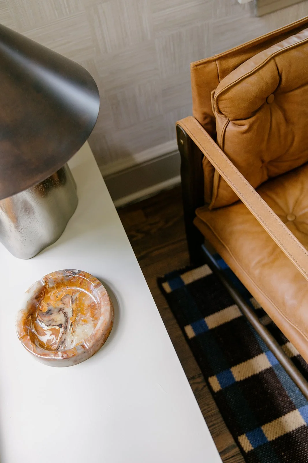 Coffee table with a decorative bowl and a lamp, adjacent to a brown leather chair with a strap resting on the armrest, next to a patterned rug.