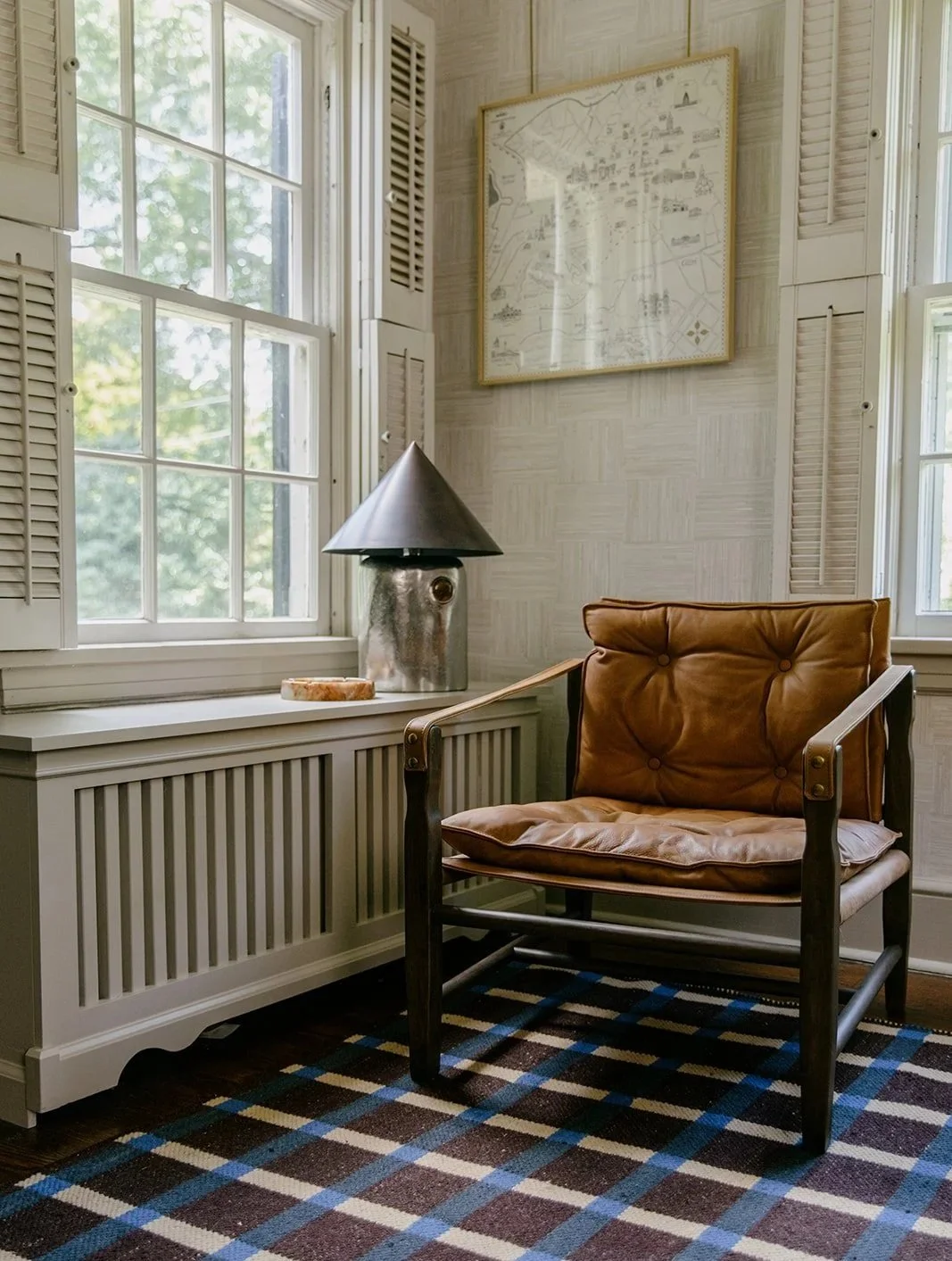 Cozy corner of a room with a brown leather armchair, a white radiator cover, a grey table lamp with a large shade, a framed map on the wall, and a checkered rug on the wooden floor near a window with open shutters.