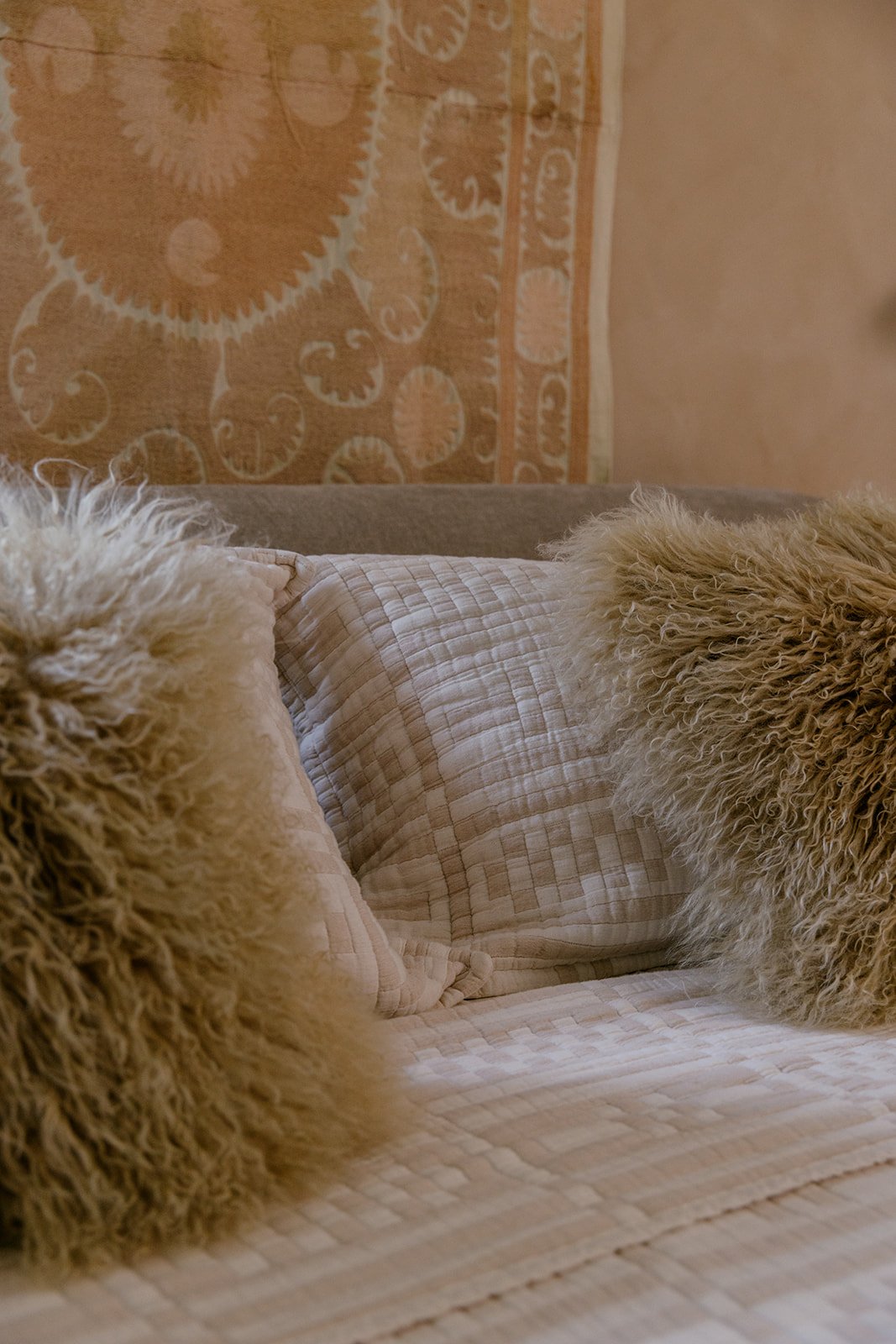 Close-up of two fluffy beige pillows on a bed with a beige checkered quilt, with a patterned rug hanging on the wall in the background.