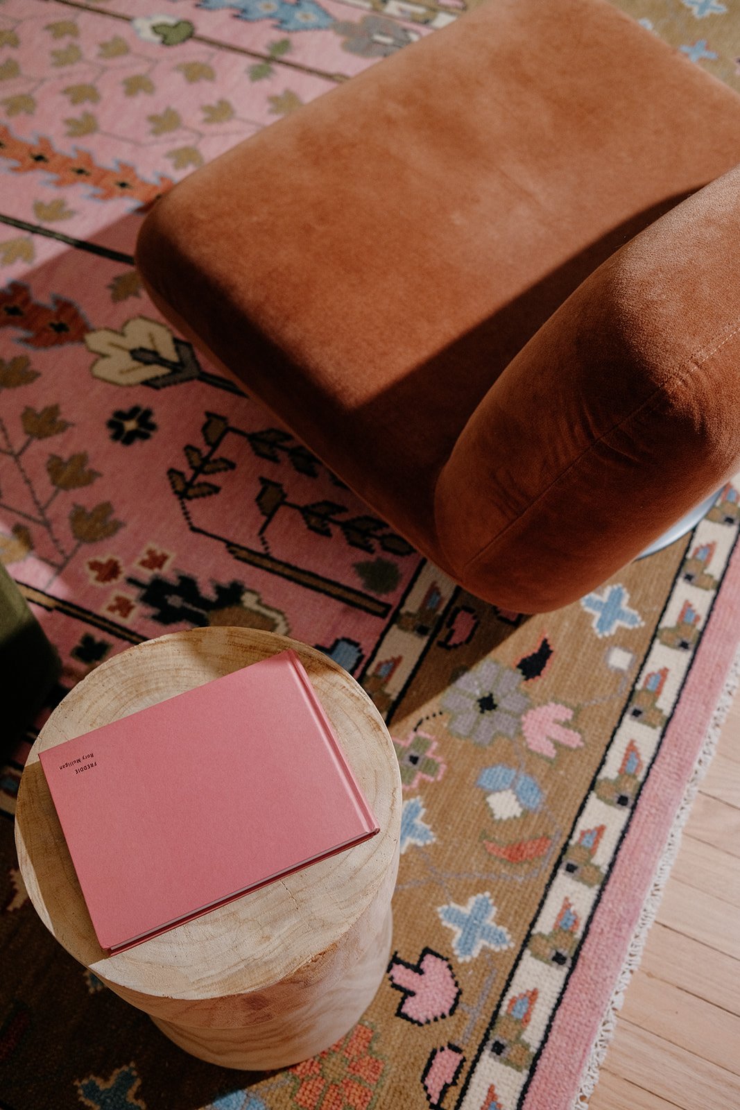 A pink book on a round wooden side table in front of a brown armchair, with a colorful patterned rug underneath.