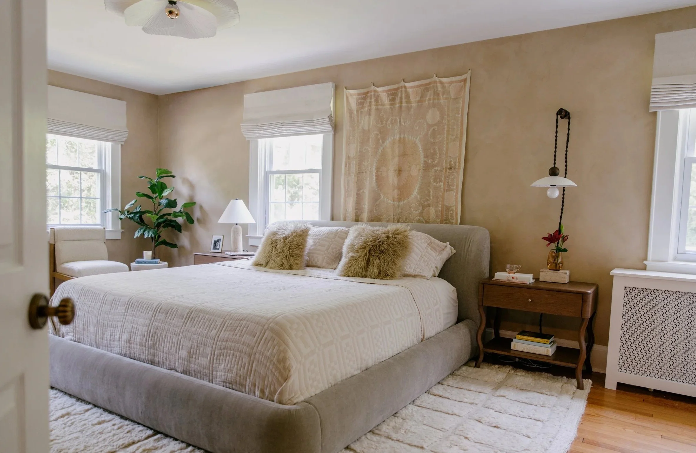A cozy bedroom with a beige bed, fluffy pillows, a nightstand with a lamp, a decorative wall hanging, and a window with blinds letting in natural light.