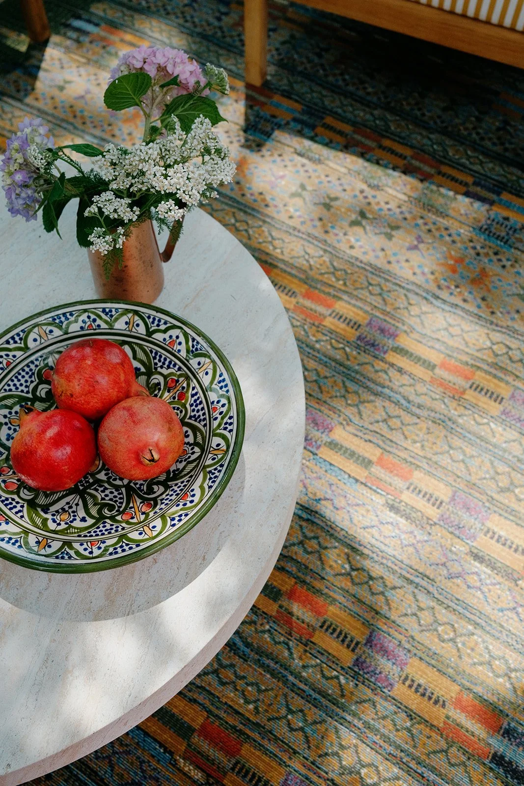 A table with three red apples on a decorative plate, a flower arrangement in a copper vase, and a colorful patterned carpet underneath.
