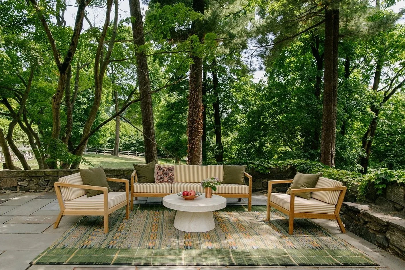 Outdoor patio with wooden furniture including a sofa and two armchairs, a round white coffee table with a bowl of apples and a flower vase, situated on a patterned green rug, surrounded by lush green trees and a stone wall.