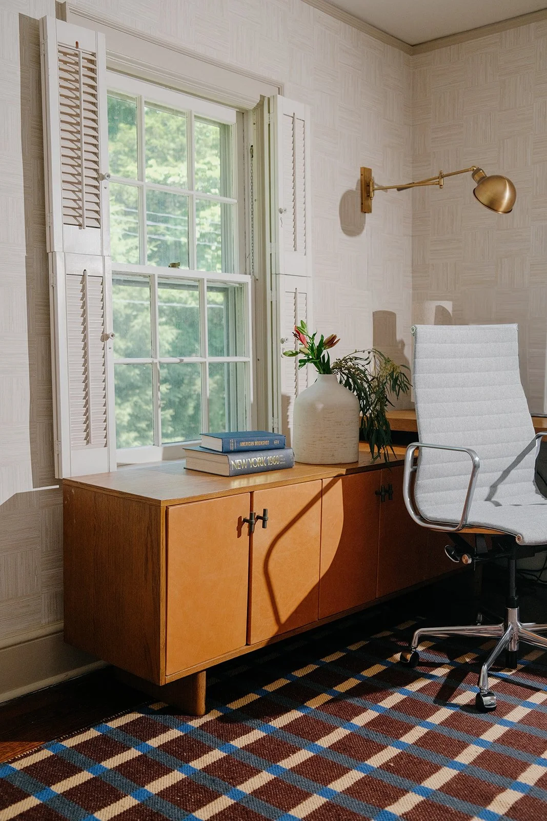 A study or office corner with a wooden sideboard, decorative books, a white vase with flowers, a white office chair, a window with shutters, a brass wall-mounted lamp, and a patterned carpet.