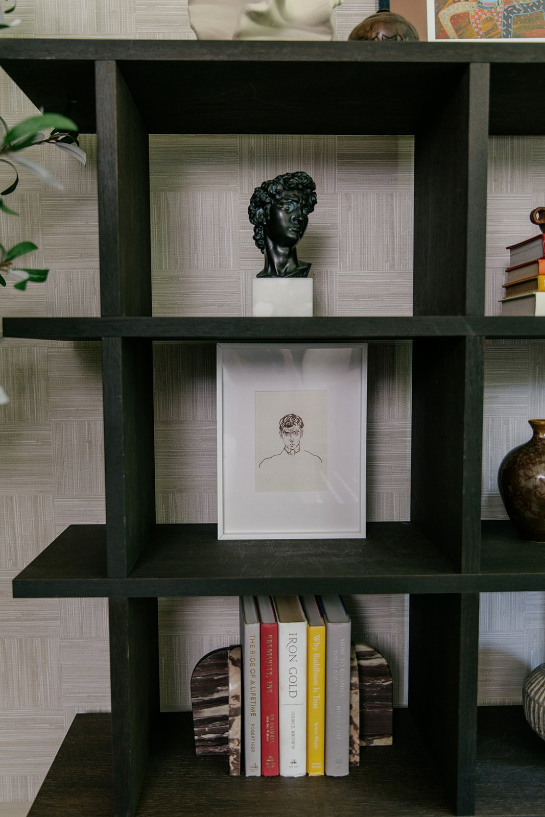 Decorative black bust sculpture on a white pedestal, framed line art drawing of a man on a shelf, books on a lower shelf, and decorative items on a wooden shelf unit.