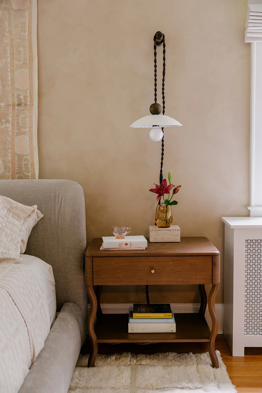 A wooden nightstand with a drawer and lower shelf sits next to a beige bed with a quilted pillow. On the nightstand, there is a clear glass bowl, a white book, and a vase with pink lilies. A vintage hanging lamp with a white shade and lightbulb is suspended above the nightstand, connected to the ceiling with a rope. The wall behind has a beige tone, with a large window partially covered by white blinds, and a floor radiator cover visible to the right.