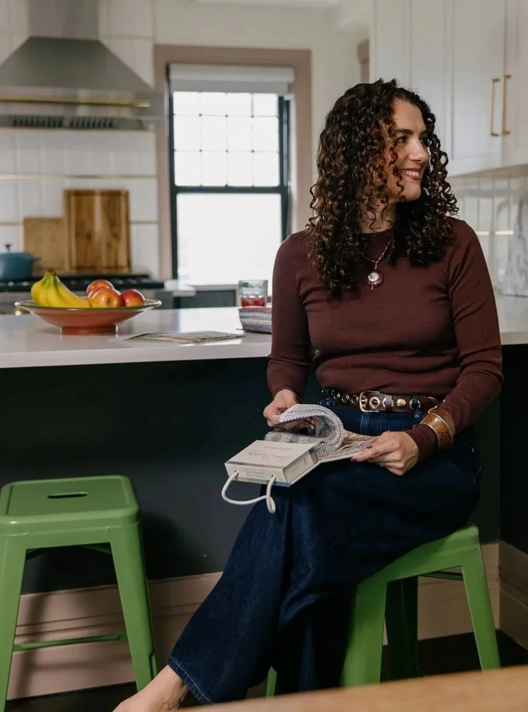 Woman with curly hair sitting on a green stool in a kitchen, holding a phone and newspaper, wearing a maroon top and blue jeans.