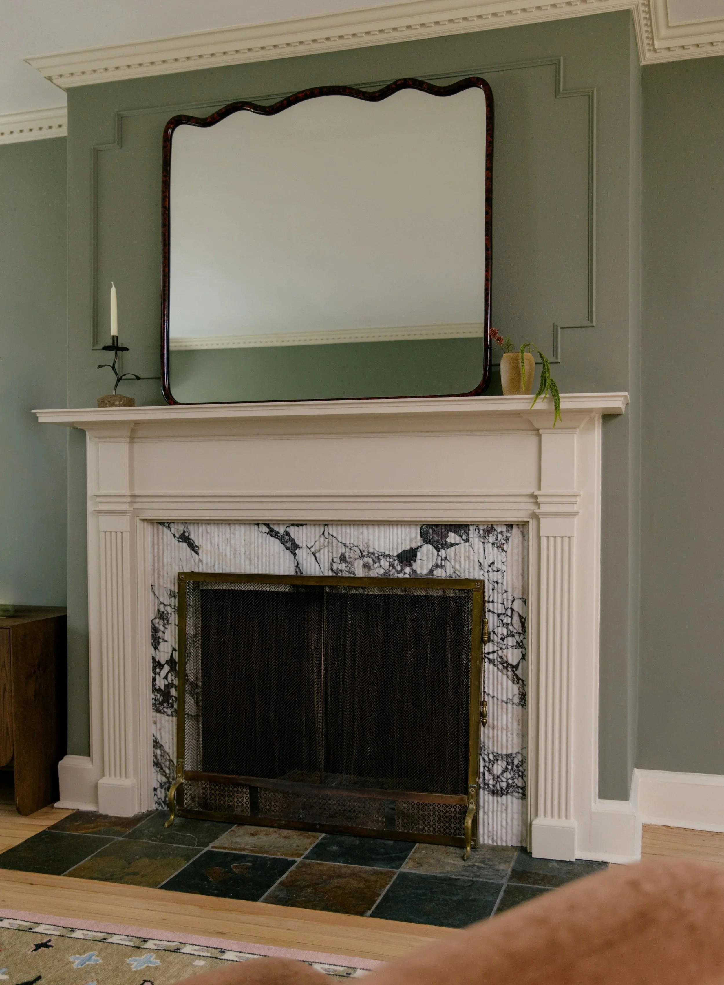 A white fireplace with a decorative mantel and black and white marble surround, a large mirror with a wavy frame above, a candle holder with a candle on the left, and a small vase with a plant on the right on the mantel.