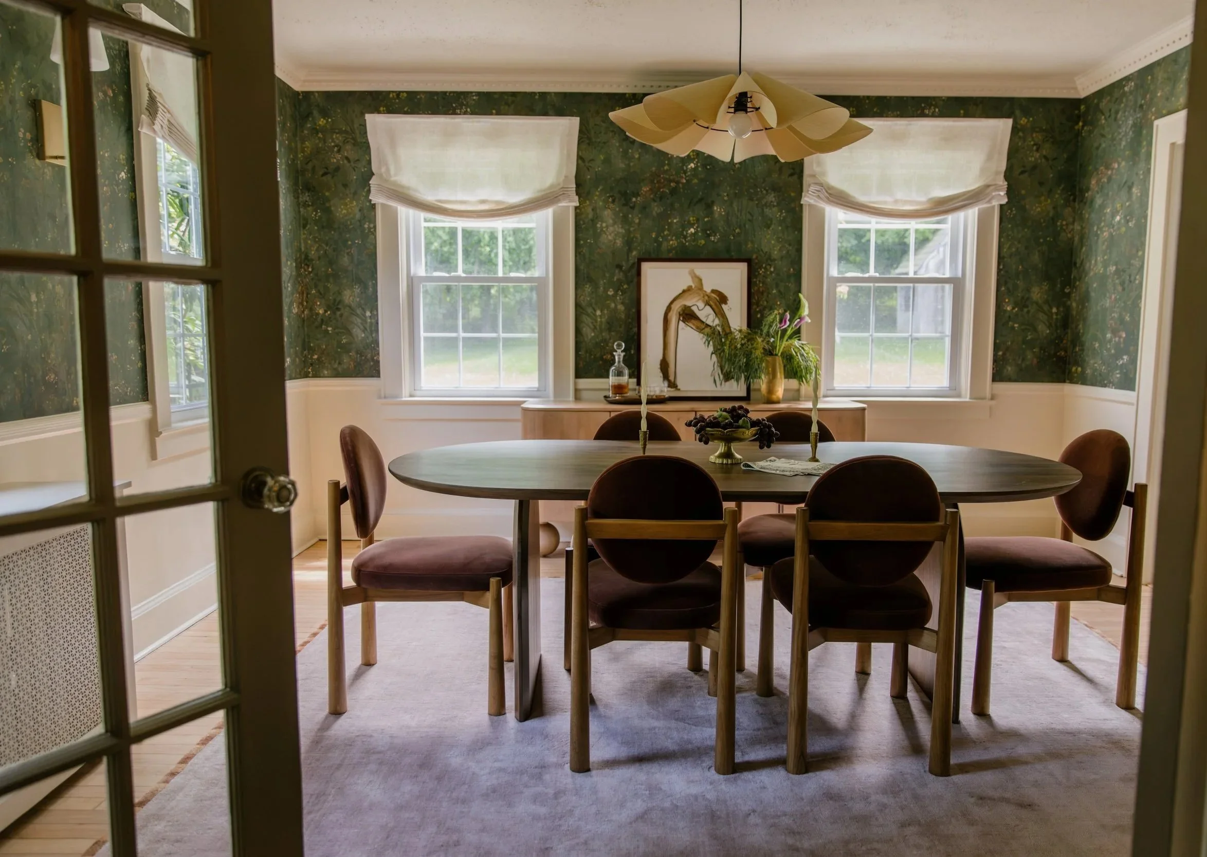 A dining room with a round wooden table surrounded by six chairs, a sideboard with a photo and plants, two large windows with white curtains, and a ceiling light fixture with a floral design. The walls are green with a floral pattern.