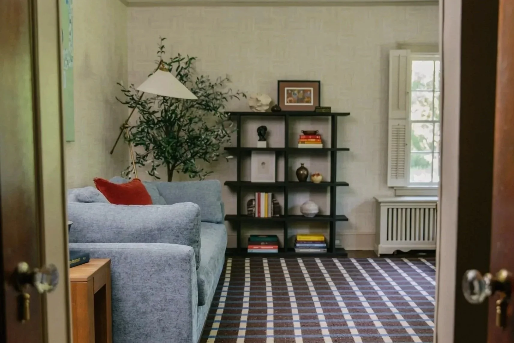Living room viewed through a doorway featuring a gray couch with a red pillow, a black shelving unit with books and decorative items, a window with white shutters, and a patterned rug on the floor.