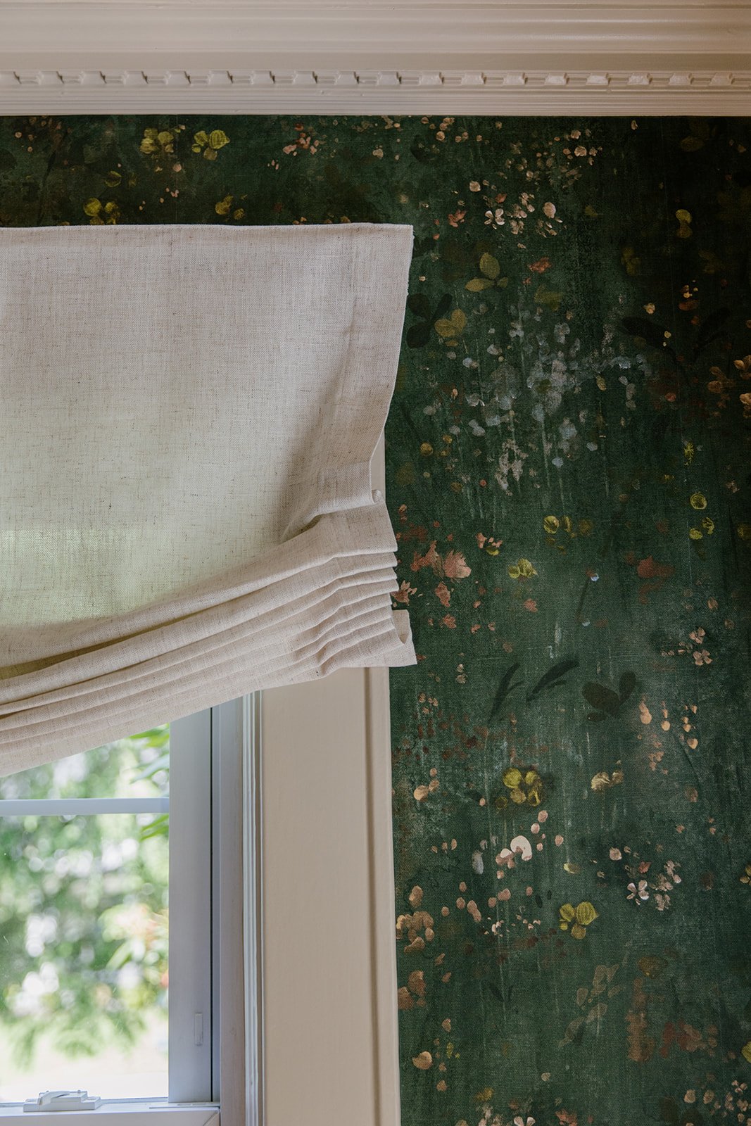 Close-up view of a window with a beige or cream-colored fabric curtain partially pulled back, revealing a green floral patterned wallpaper or wall behind it.