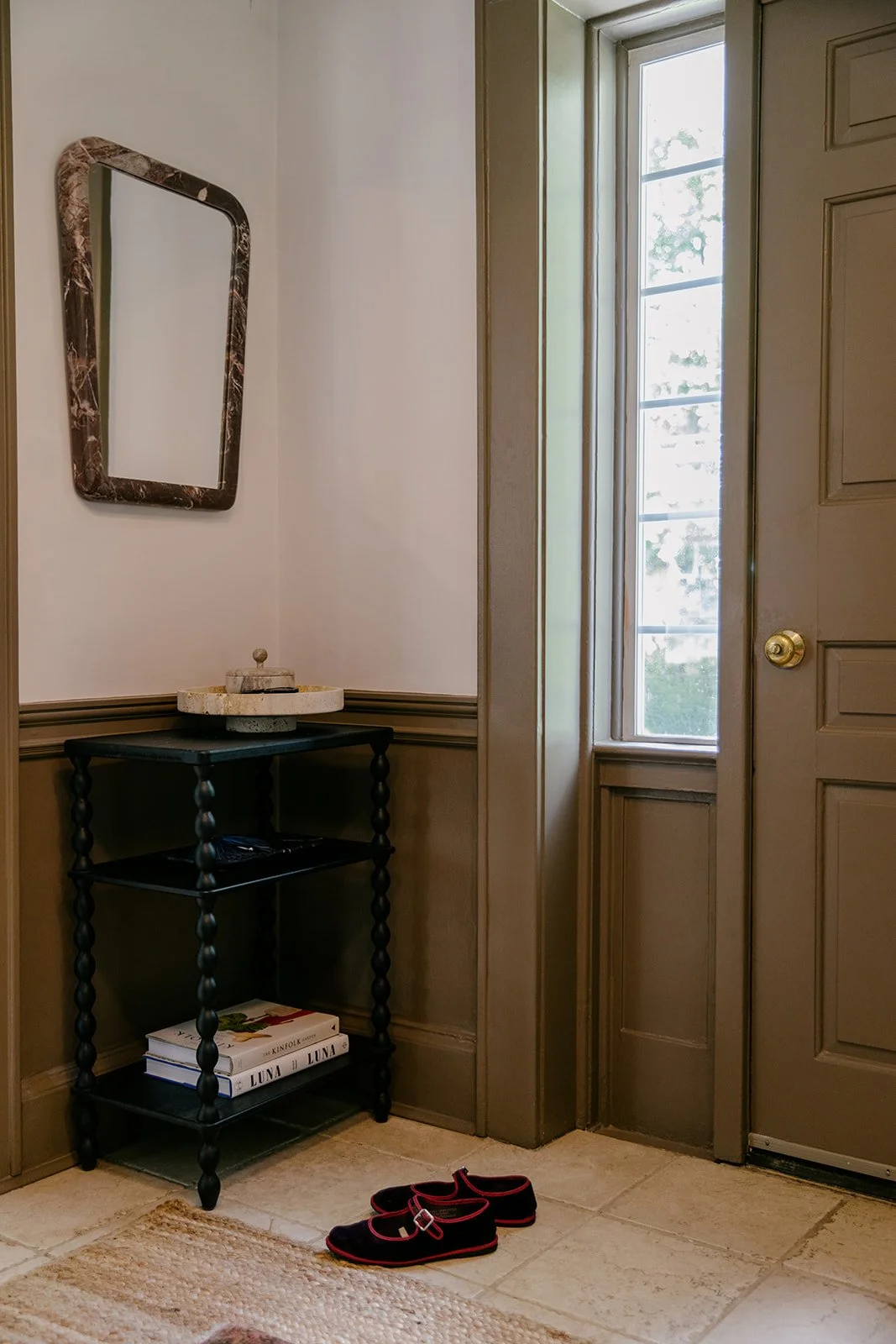 Interior of a house entryway with a black console table holding a decorative bowl and some books, a wall mirror with a marble frame, a window, a door with a gold doorknob, and a pair of black slippers with pink trim on the beige tiled floor.